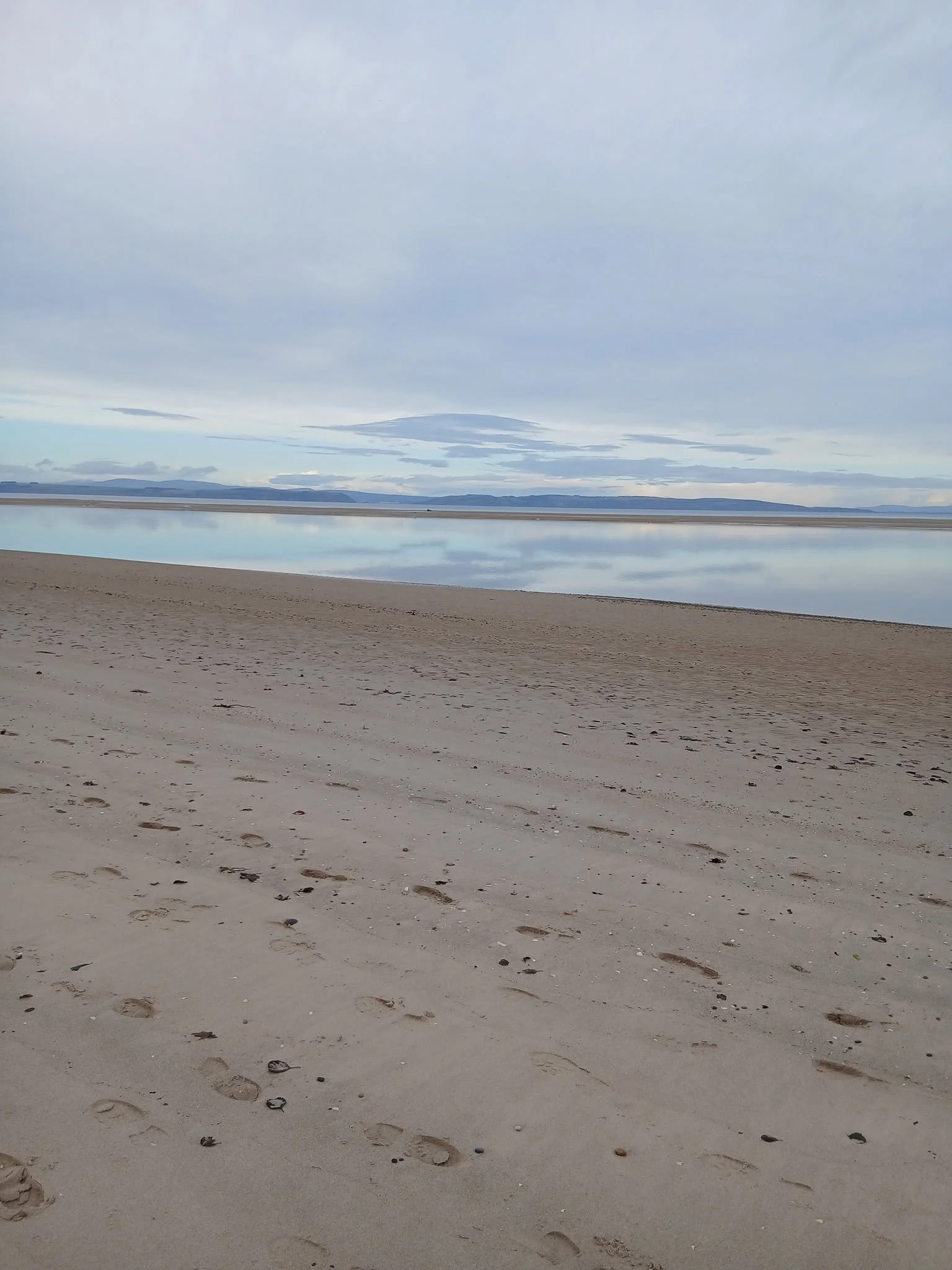 A sandy beach with footprints leading towards calm water, with distant hills under a cloudy sky.