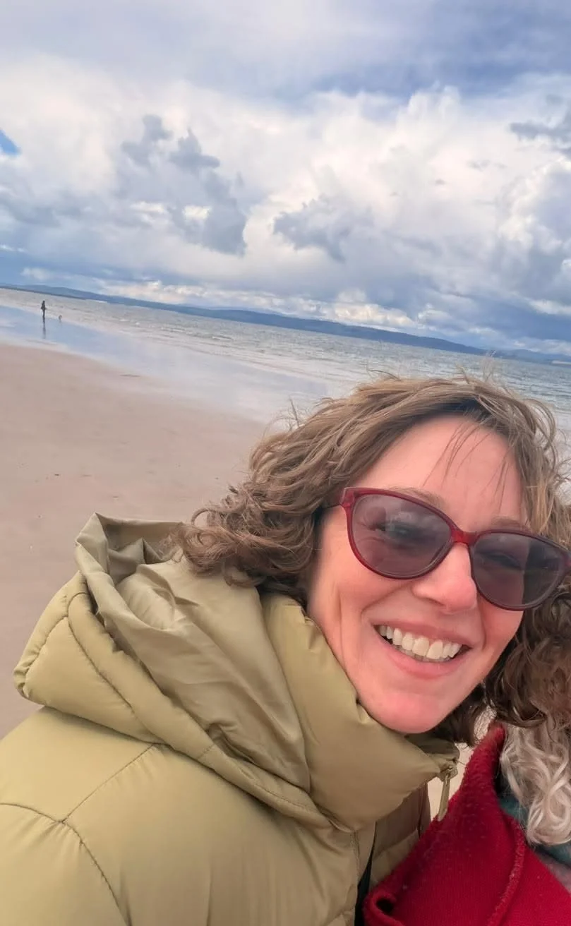 Smiling woman with sunglasses taking a selfie on a beach with cloudy sky and ocean in the background.