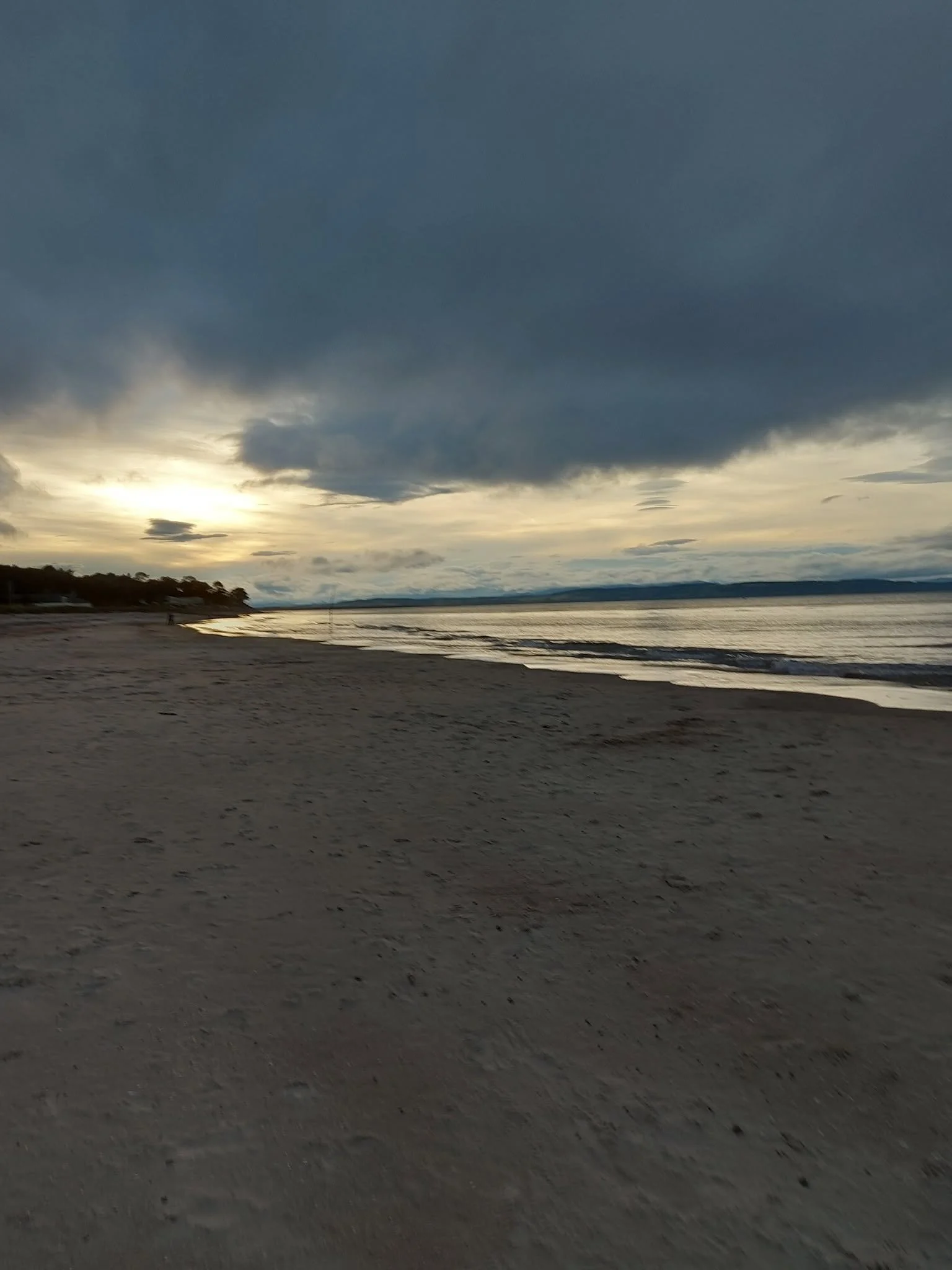 A beach scene at sunset with dark clouds overhead, calm water, and a sandy shore in the foreground.