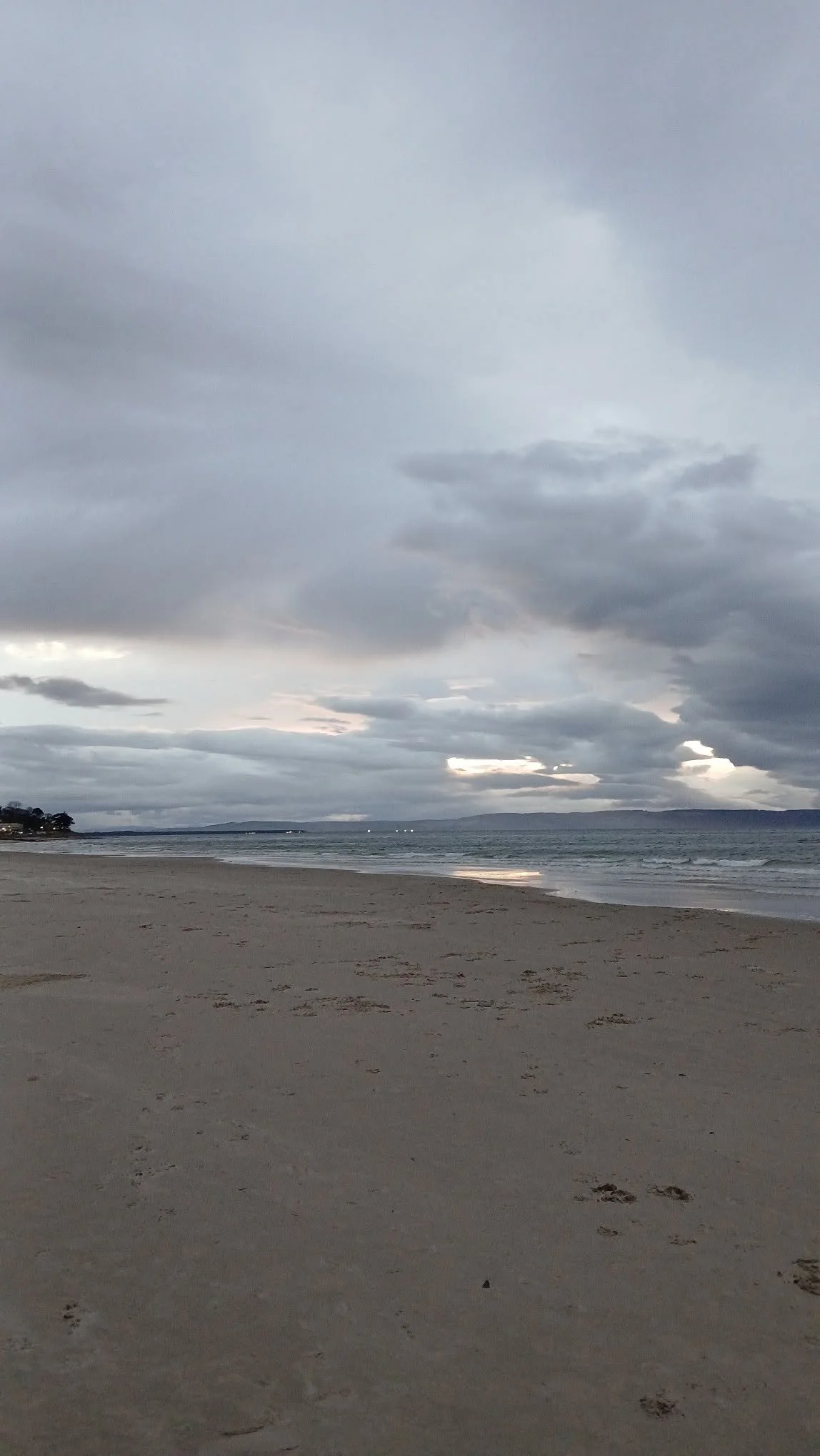 A cloudy beach scene with sandy shore, ocean waves, and a cloudy sky over distant land.