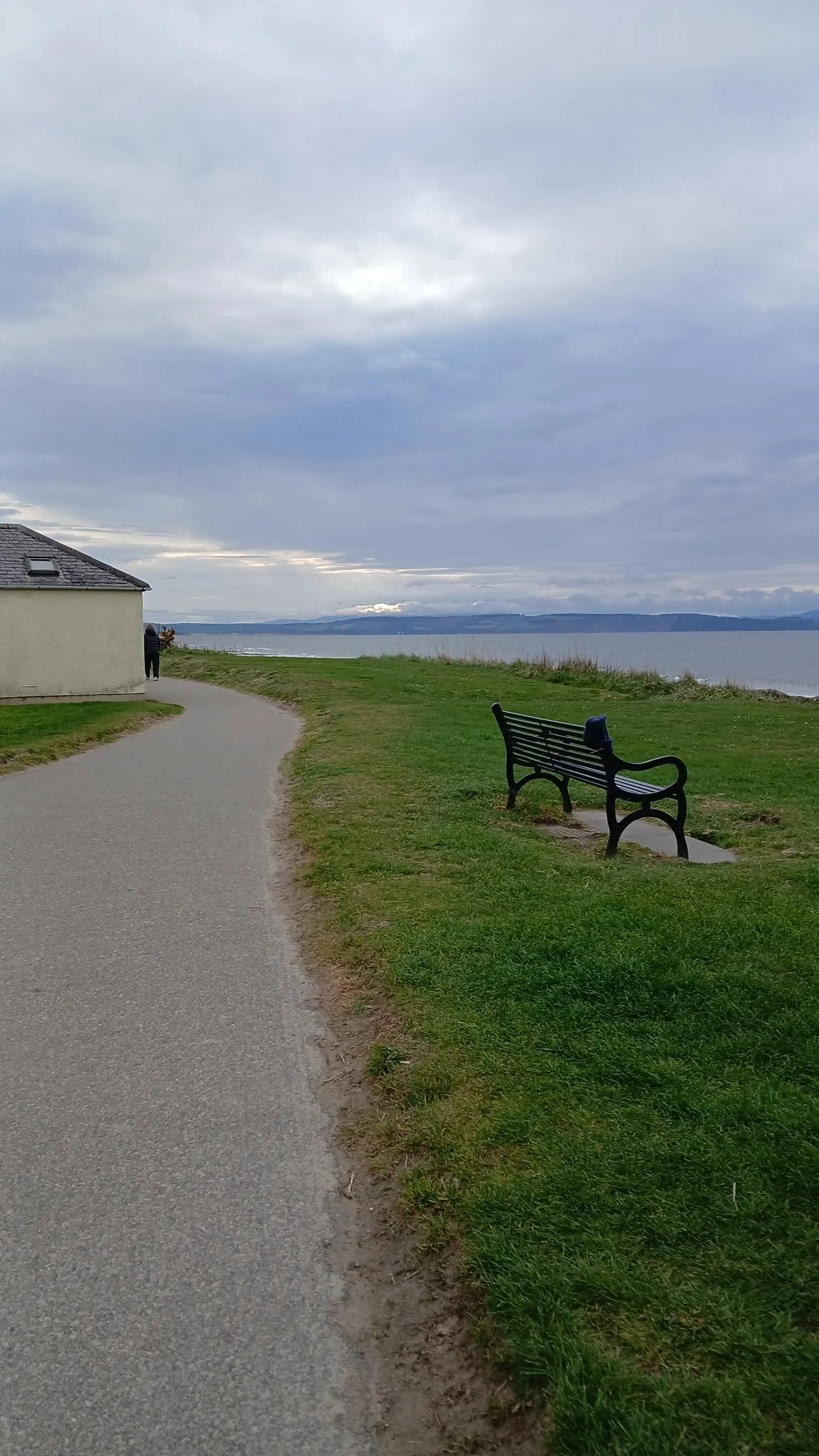 A pathway along a grassy area next to a waterfront with a bench, a small building, and a person looking out at the water under a cloudy sky.