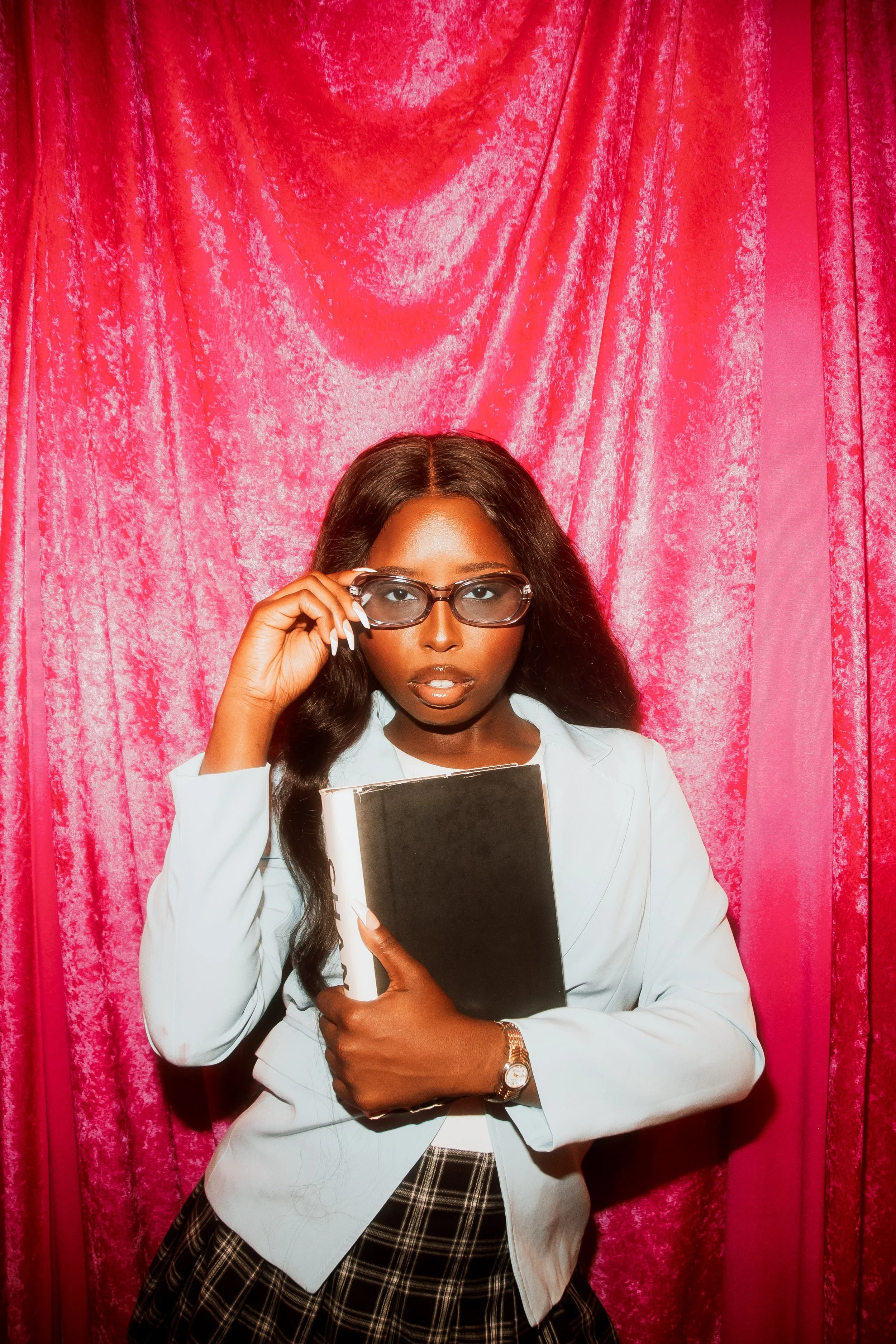 A woman with dark skin, wavy hair, wearing glasses, a white blazer, patterned skirt, holding a black book, standing in front of pink curtains.