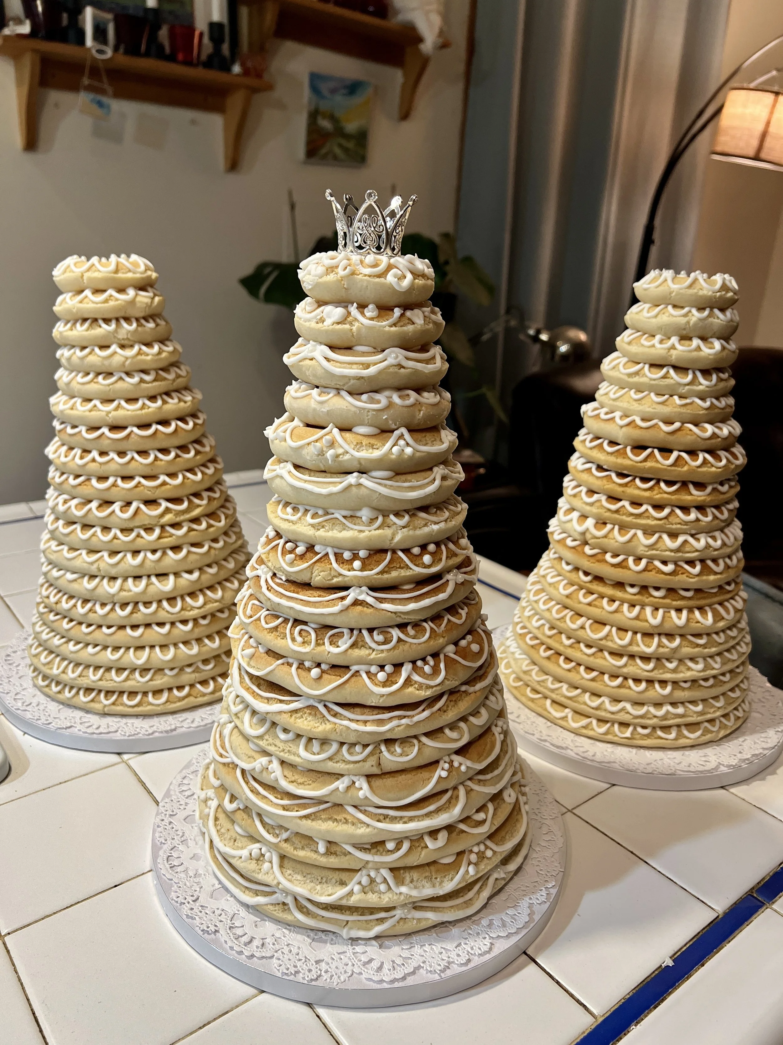 Three Kransekake arranged on round white doilies with intricate patterns, featuring white icing designs. One stack has a small silver crown on top. The background shows a kitchen or dining area with shelves, artwork, and a lamp.