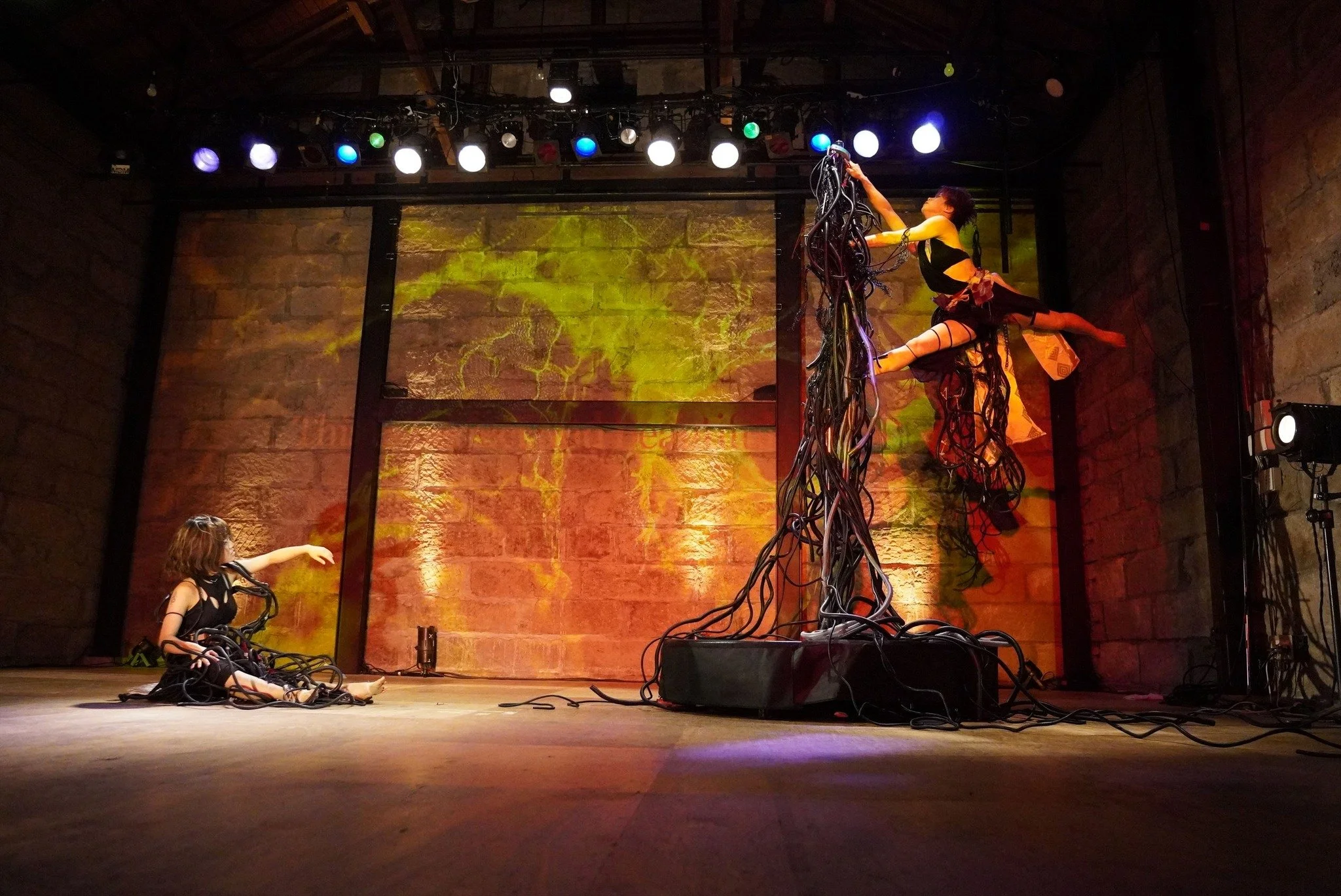 A stage performance featuring dancers with wires and cables. The dancers are in dynamic poses, with one lifting her leg high and another sitting on the stage. The stage has colorful overhead lighting and a brick wall background.