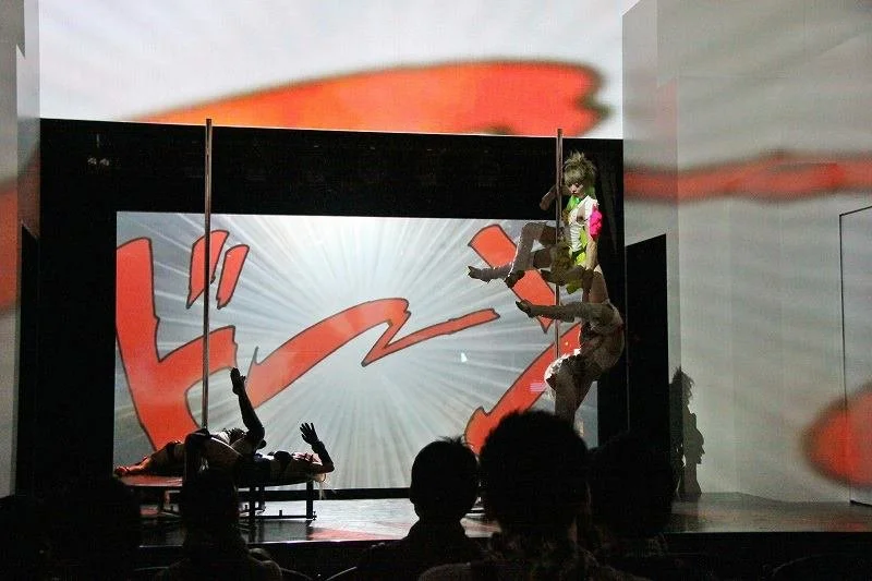 A stage performance with clown or character jumping or falling in front of a large superhero logo backdrop, with an actor in colorful costume and audience silhouettes in the foreground.
