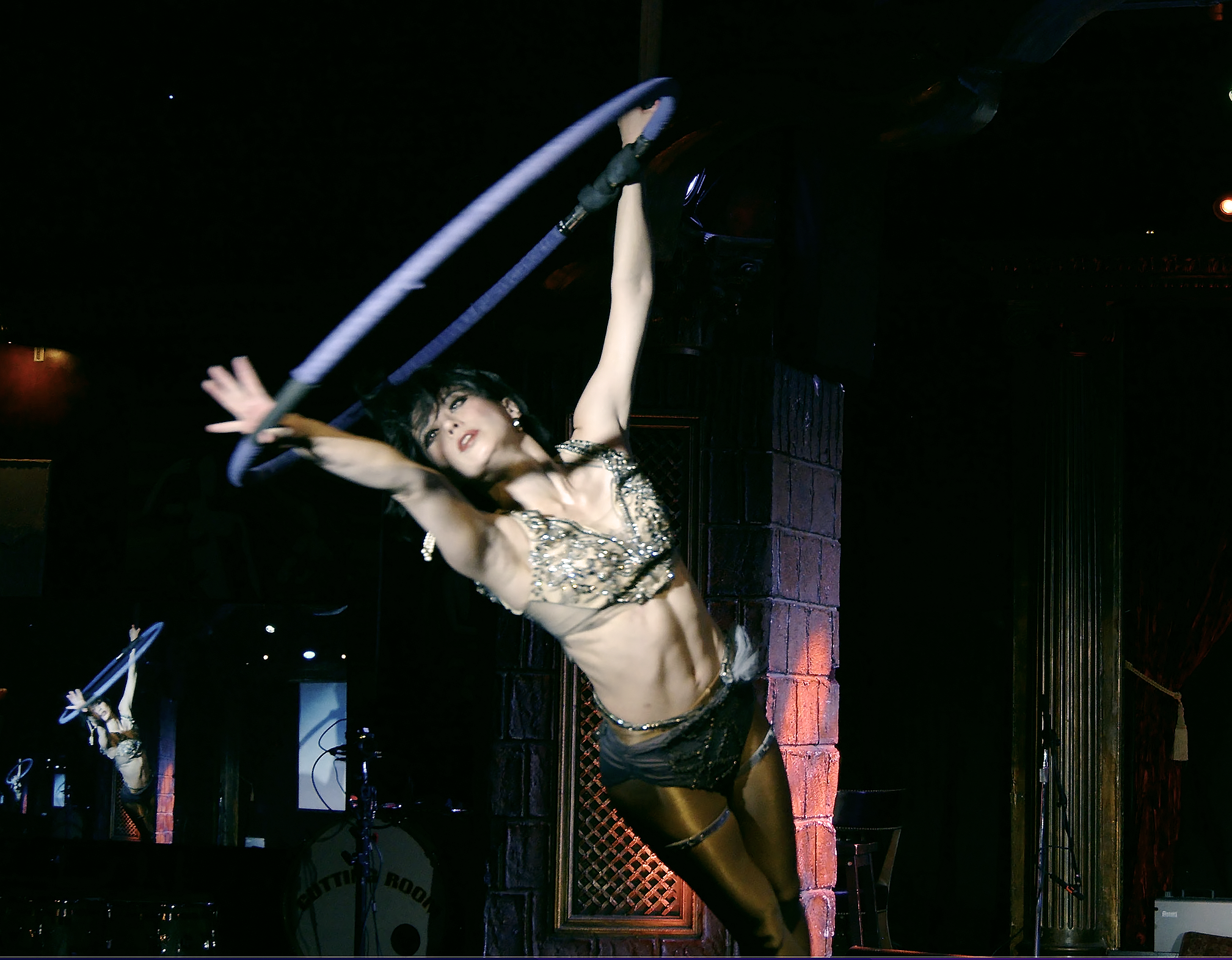 A woman dressed in burlesque attire dancing on stage, with a brick wall and dark background, performing under dim lighting.