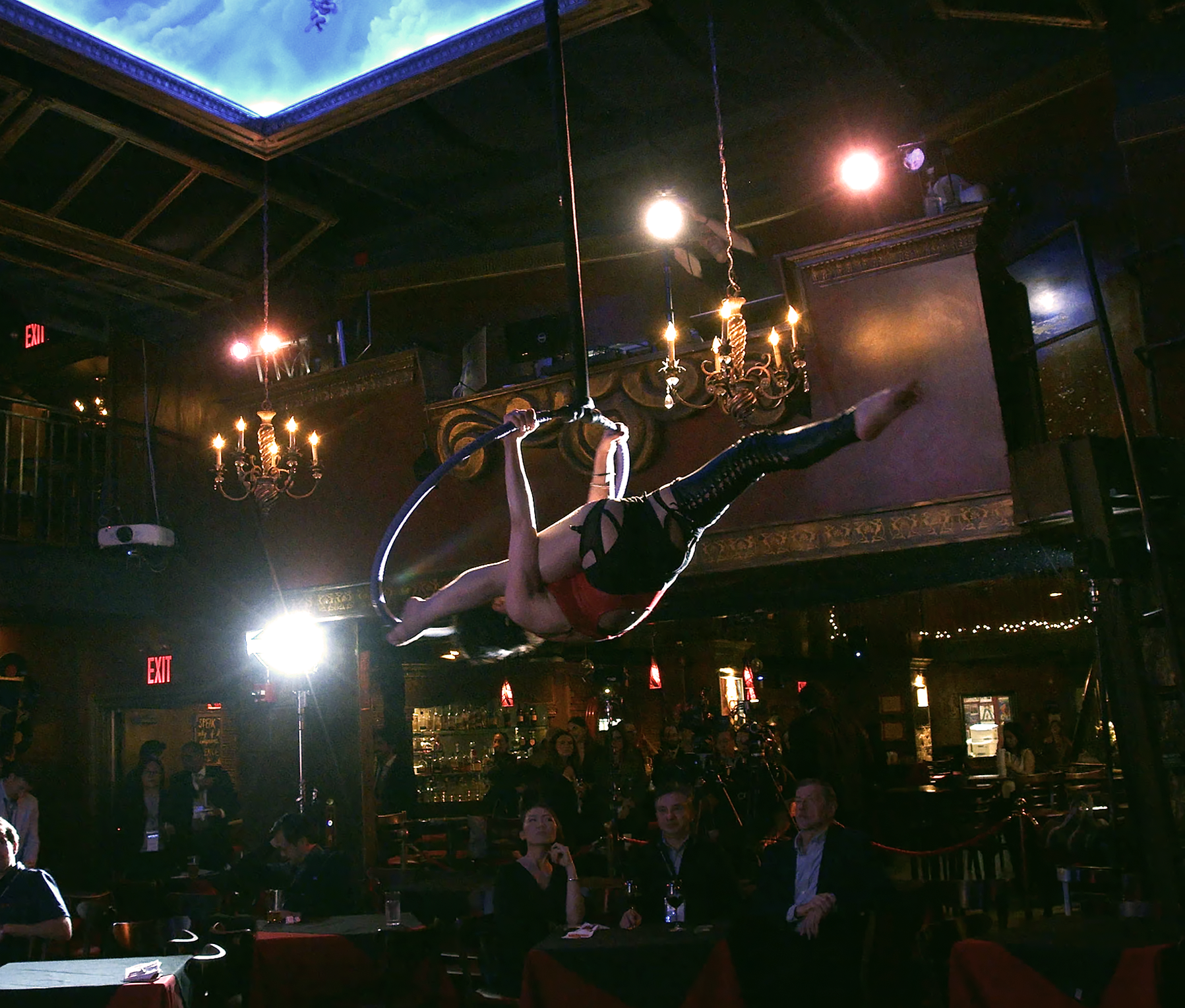 A woman performing aerial silk acrobatics hanging upside down inside a dimly lit restaurant or bar. The ceiling has decorative chandeliers, and there are people seated at tables watching the performance.