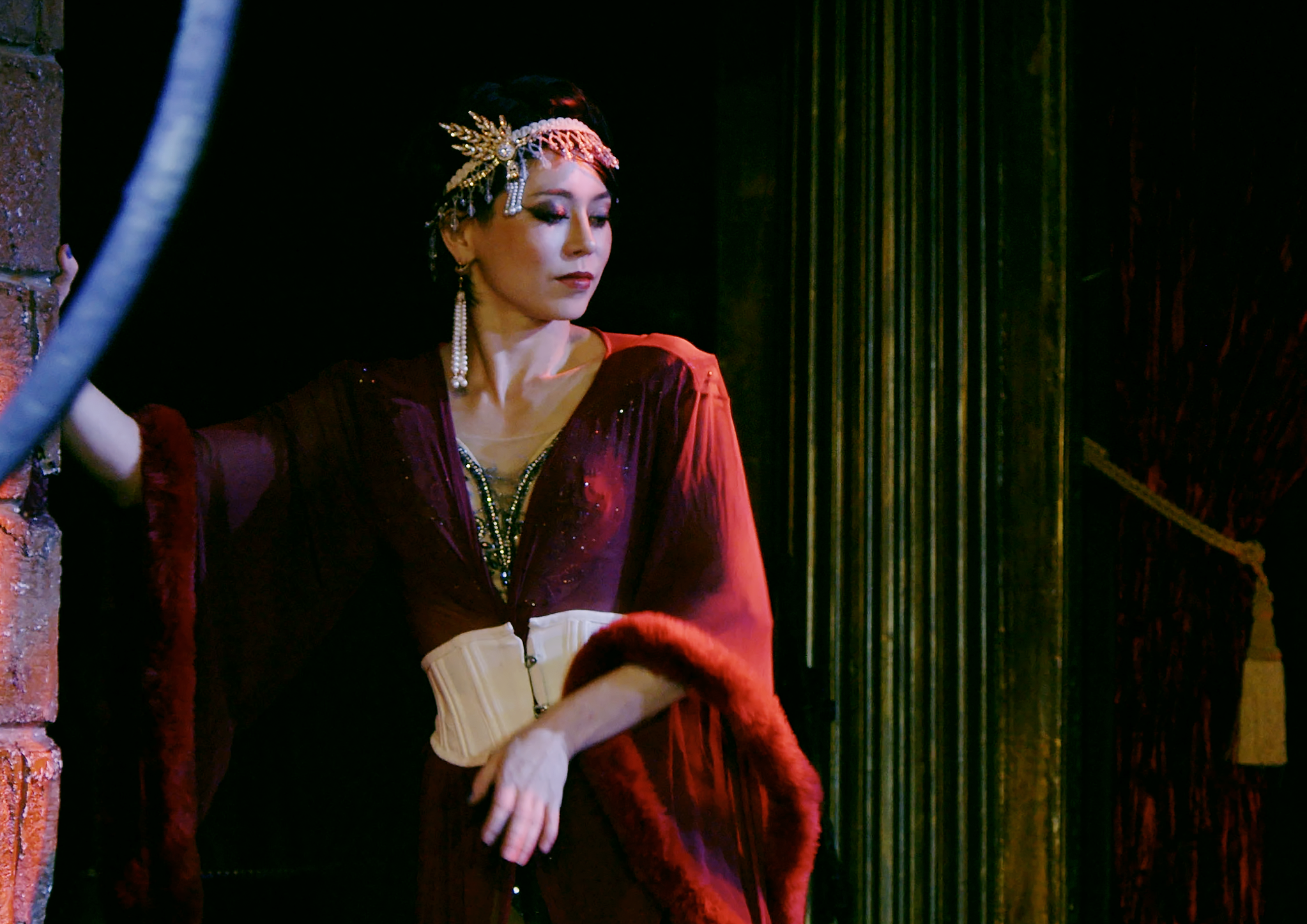 A woman dressed in vintage or costume attire, with elaborate jewelry, wearing a regal-looking outfit, standing against a dark background with curtains.
