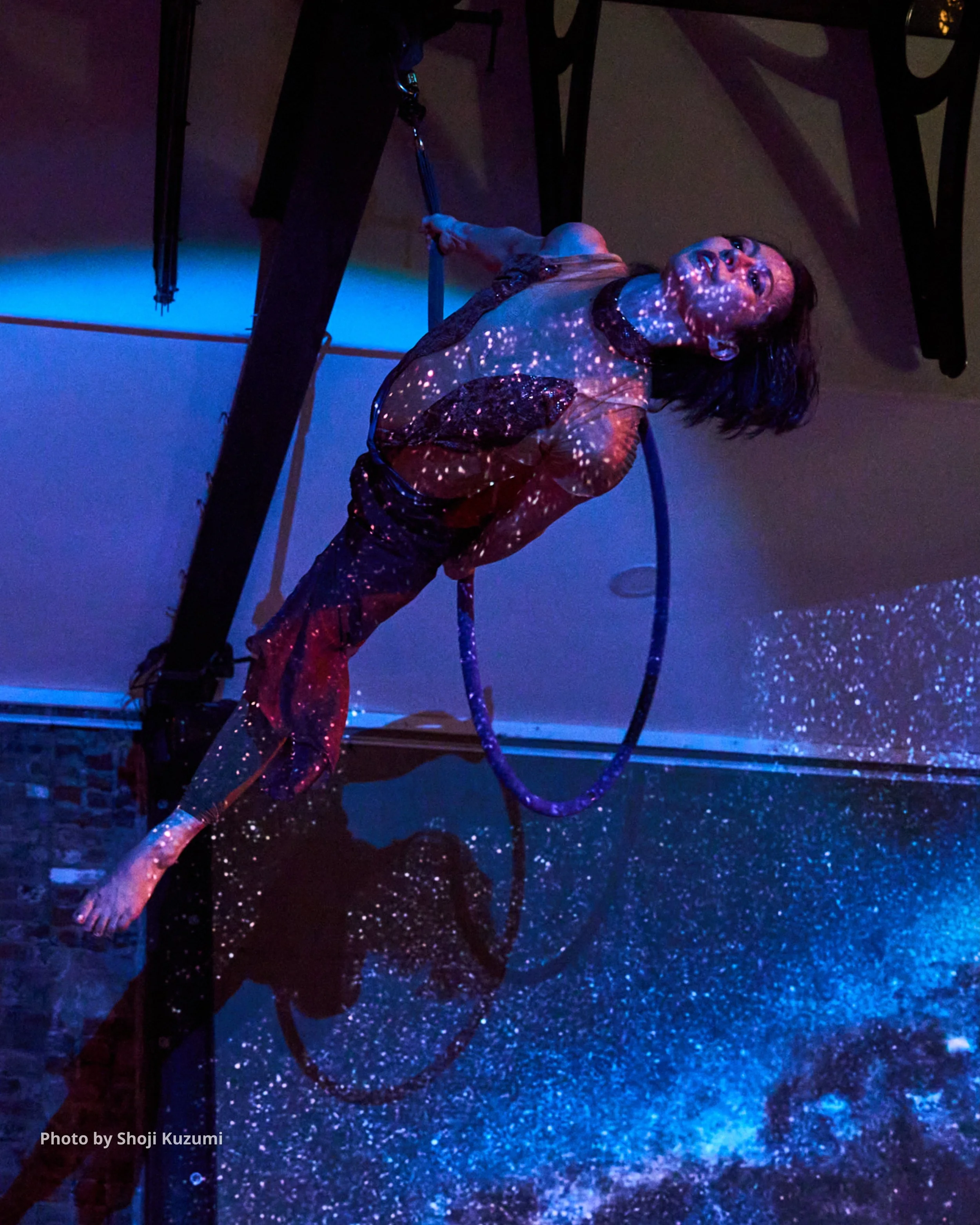 A person appears to be in space or cosmic environment, with projected stars on their face and clothing, standing in a dark room with neon lighting, holding a hula hoop. The scene has a surreal, artistic atmosphere.