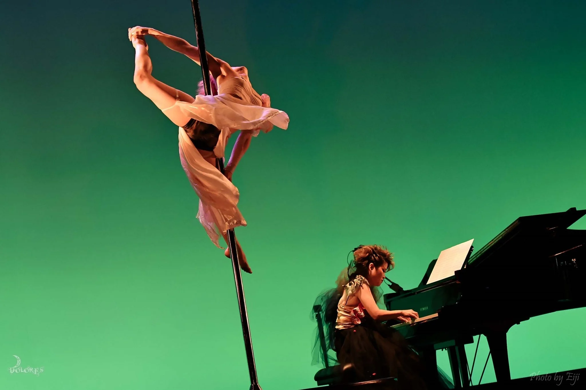 Musicians performing on stage with a green background, featuring a woman playing the piano and another person dancing or performing aerial acrobatics.