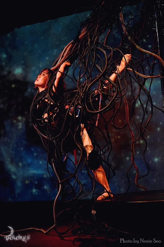 A woman dressed in a black costume with long black cables tangled around her, holding onto a large structure above her head, standing on a stage with a galaxy-themed background.