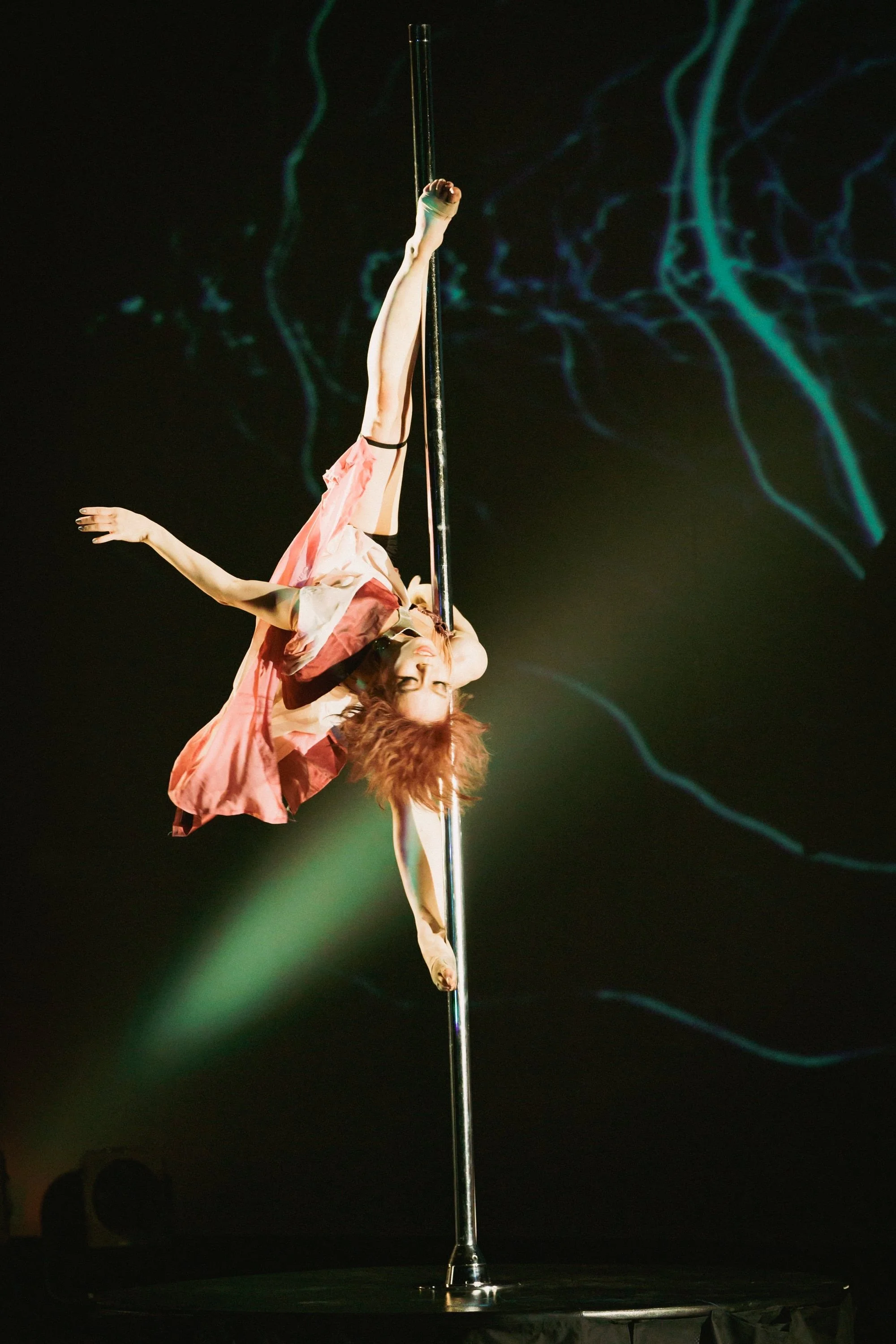 A female performer with curly hair, dressed in a pink and beige outfit, is performing an aerial act on a vertical pole during a stage show. The background is dark with blue and green lighting effects.