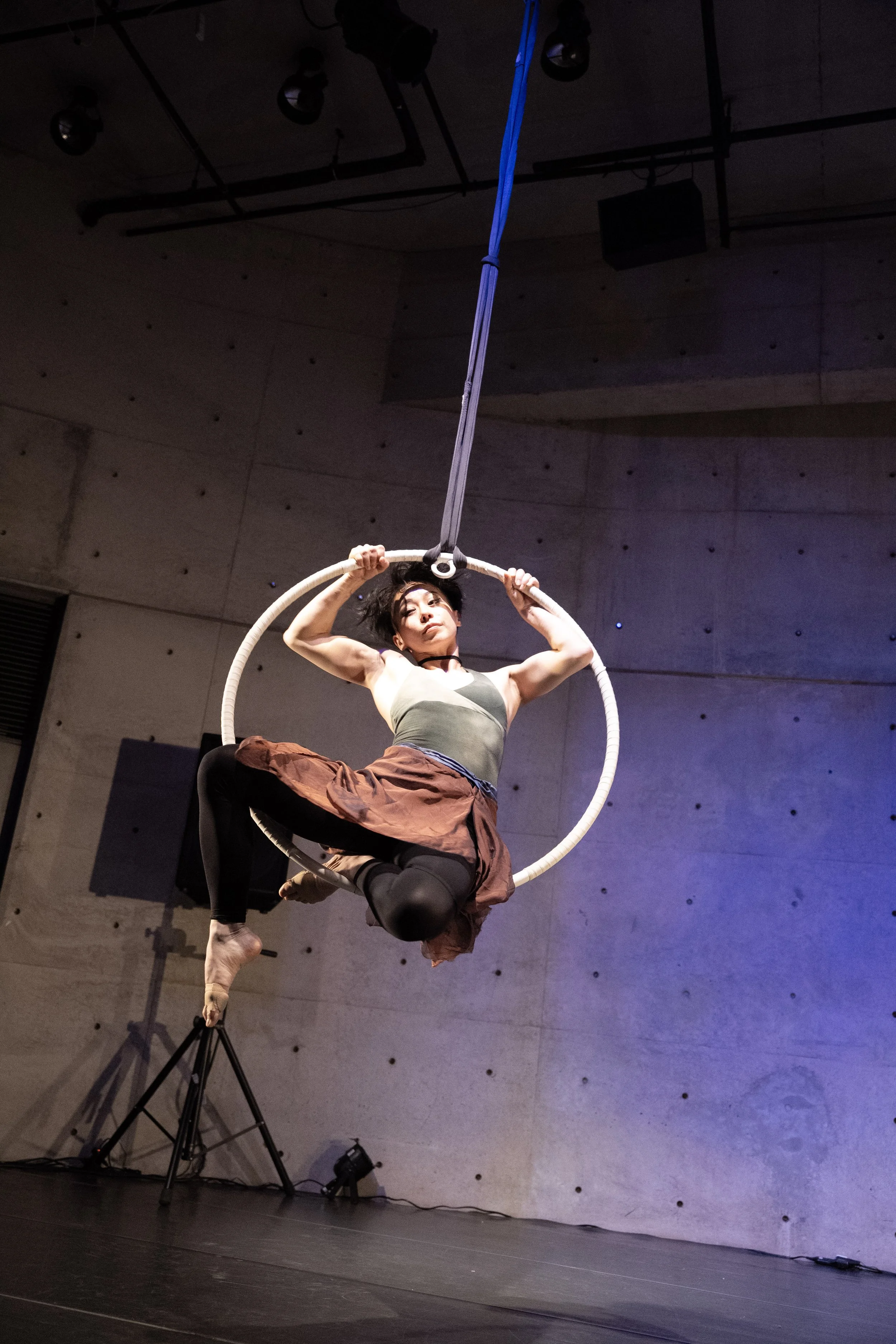 A woman practicing aerial acrobatics on a hoop, hanging from the ceiling in a dark indoor studio with a concrete wall background.