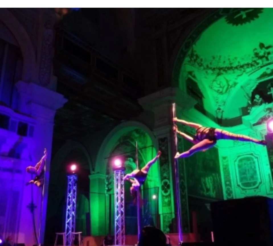 Three female acrobats performing aerial pole routines on indoor stage with colorful lighting and ornate architecture.