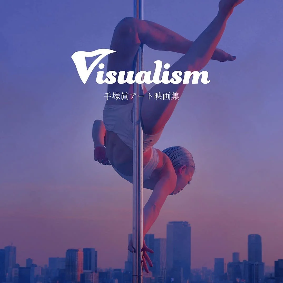 A person performing a pole dance or acrobatic move on a pole against a city skyline at sunset with purple and pink sky, with the word 'Visualism' and Japanese characters overlayed.