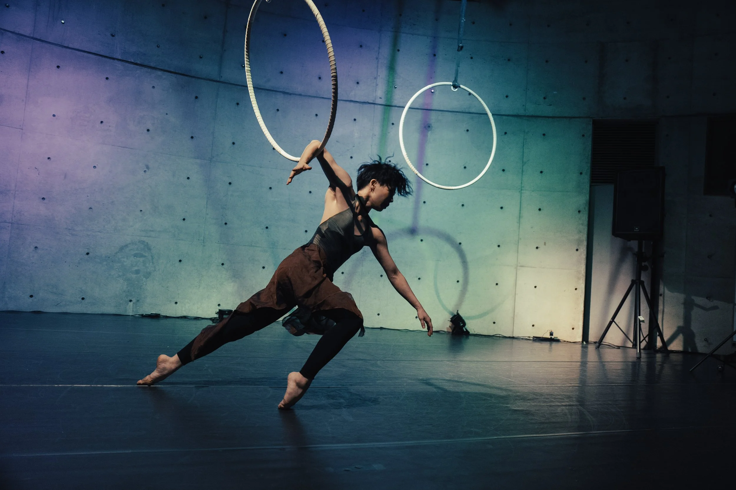 A dancer performing on stage with large aerial hoops hanging from the ceiling, in a dimly lit room with a plain concrete wall background.