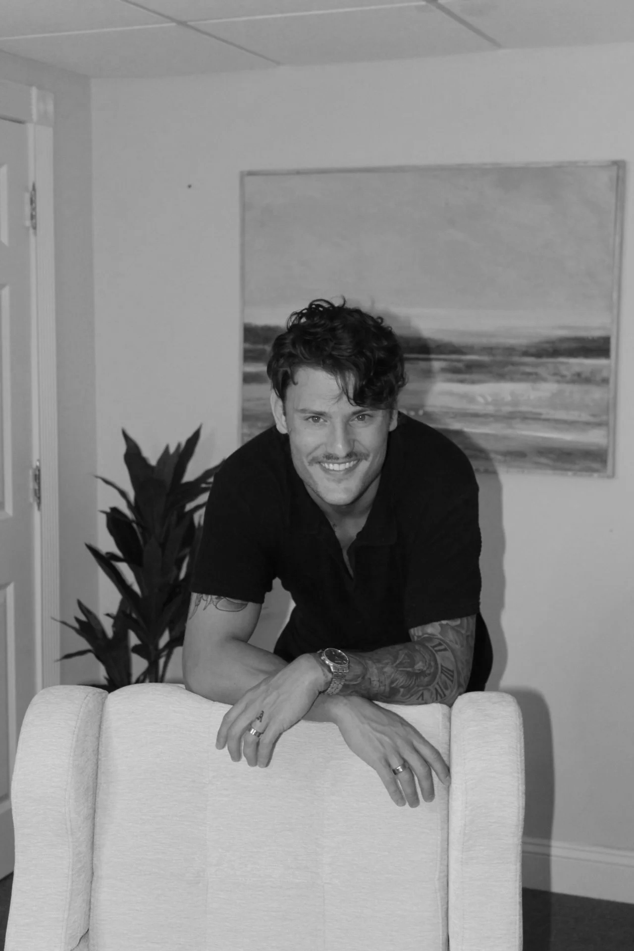 A smiling man with tattoos on his arms, wearing a black shirt and a wristwatch, leaning on the back of a light-colored armchair in a room with a landscape painting, a potted plant, and a door in the background.