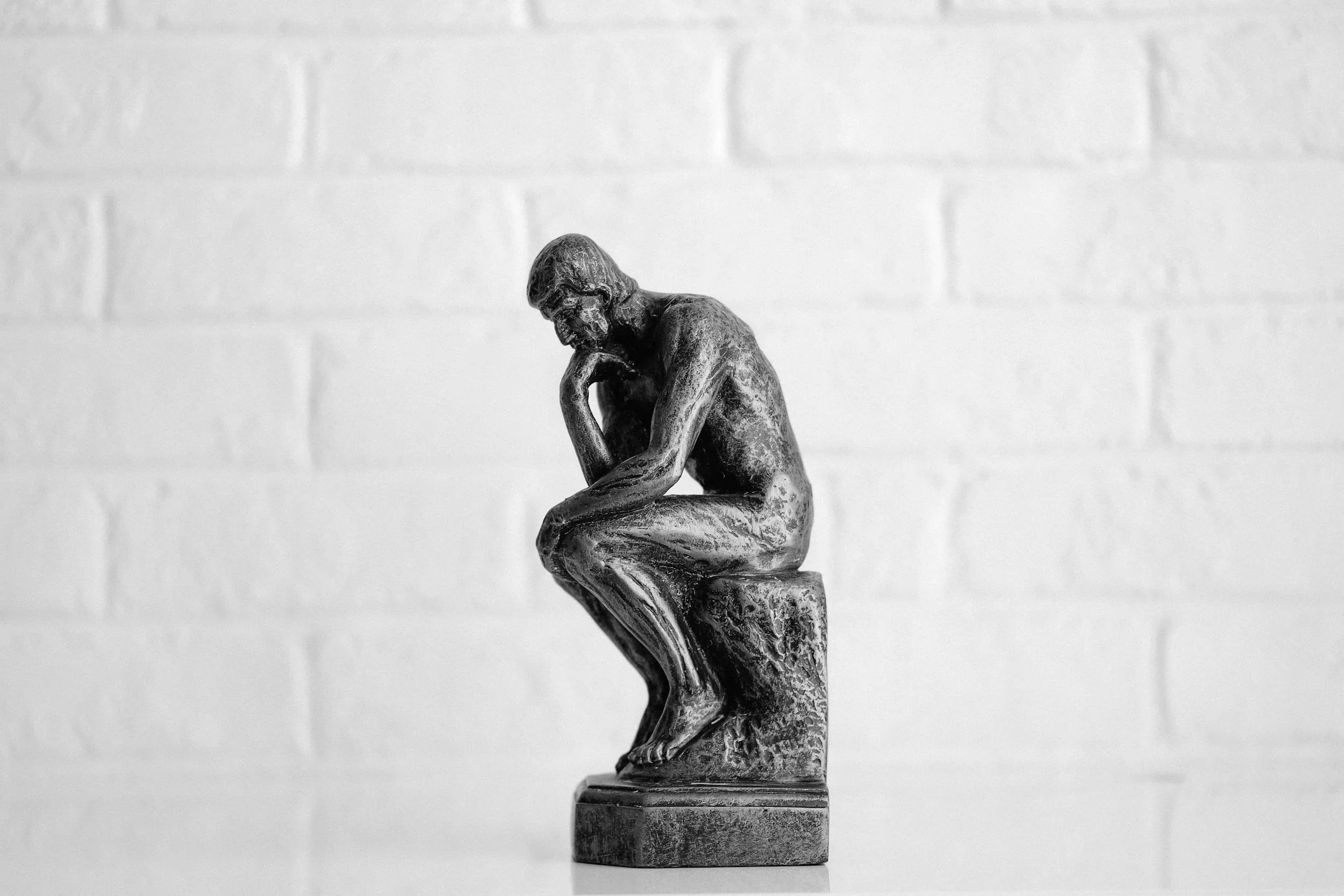 A metallic sculpture of a person sitting with their head resting on their hand, set against a plain white wall.