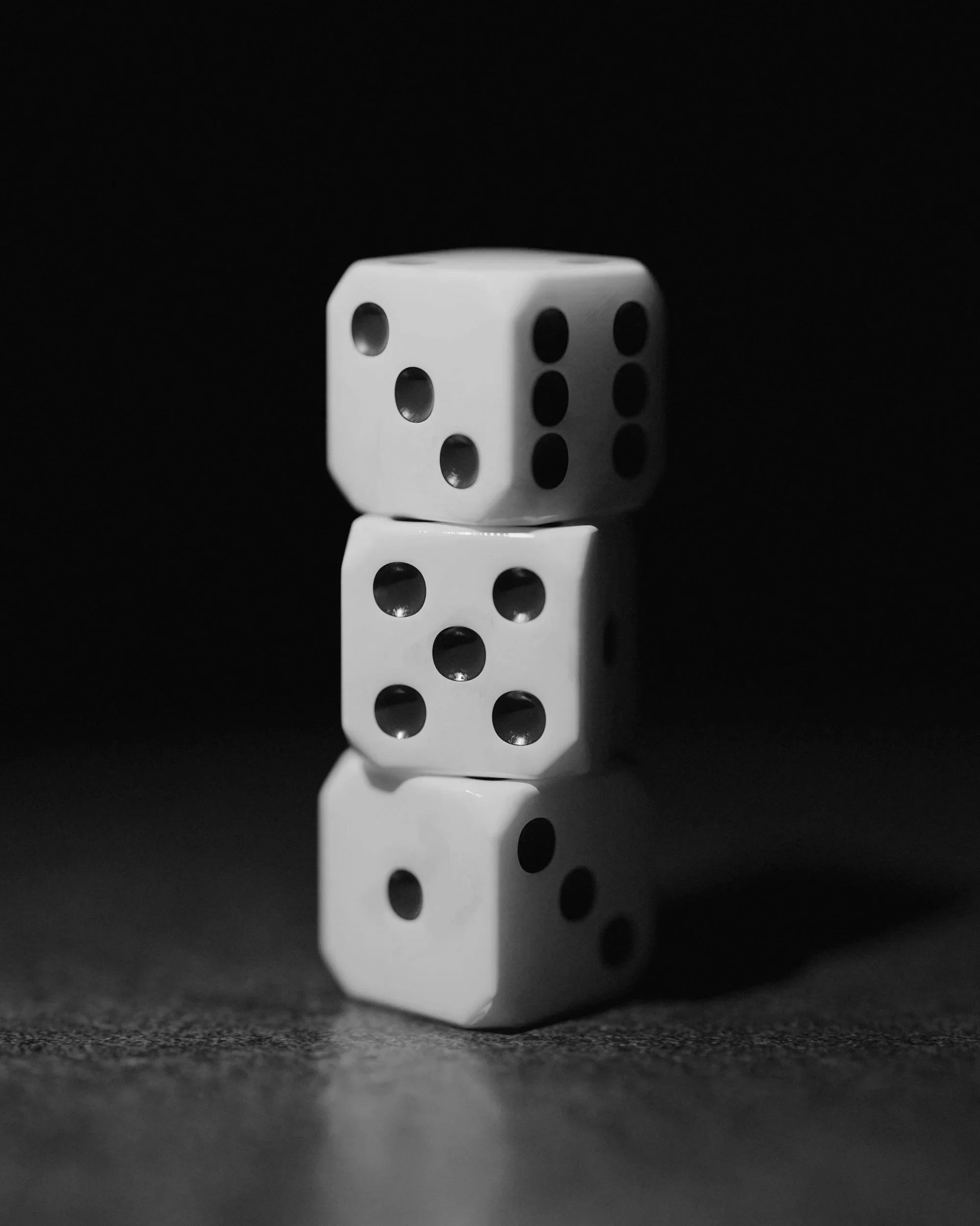 Four white dice with black dots stacked vertically on a dark surface against a black background, with the current perspective showing the 3, 5, 4, and 6 sides from top to bottom.