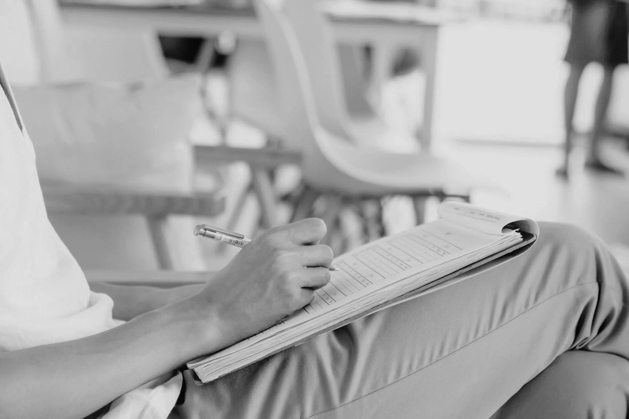 A person wearing a white shirt is holding a pen and writing on a notepad. The notepad is resting on their lap, and they are seated in a classroom or meeting room with blurred chairs and tables in the background.