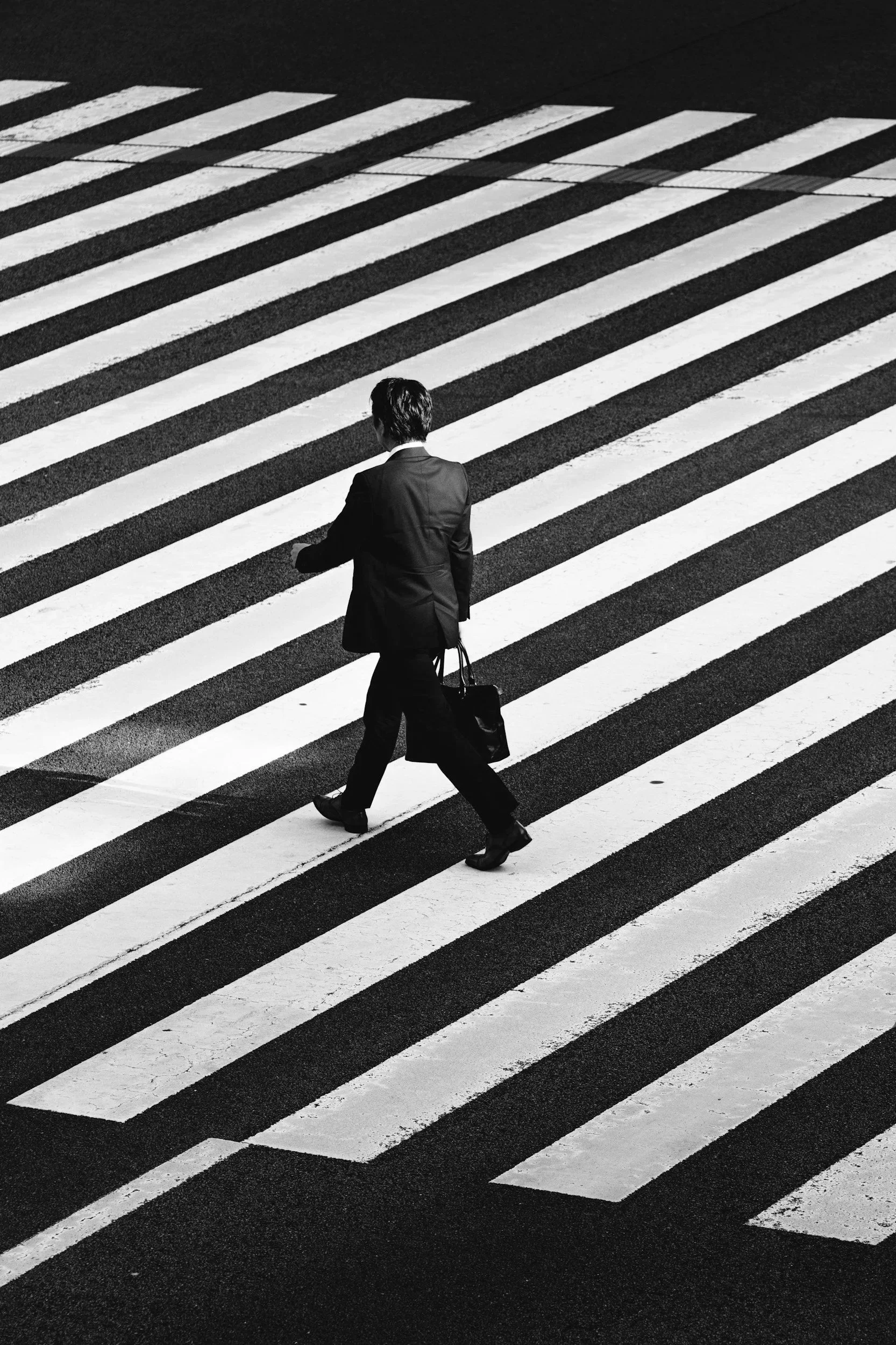 Black and white photo of a person in business attire crossing a city street at a crosswalk.