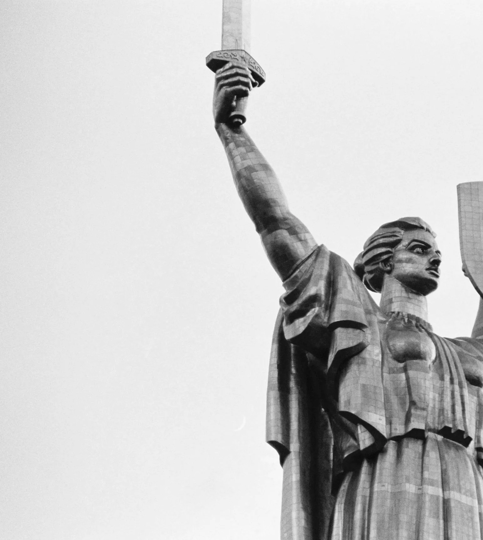 Black and white photograph of the statue of liberty, showing her face, raised arm holding the torch, and the robe she is wearing.