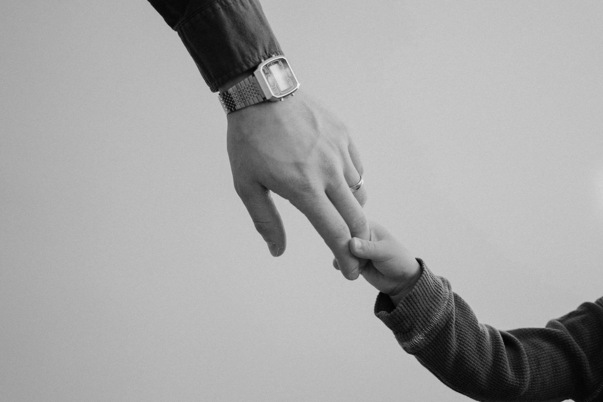 A close-up of a person wearing a wristwatch reaching out to hold a child's hand. The person is dressed in a dark long-sleeve shirt or jacket, and the child's hand is smaller and wearing a long-sleeve shirt. The background is plain and light-colored.