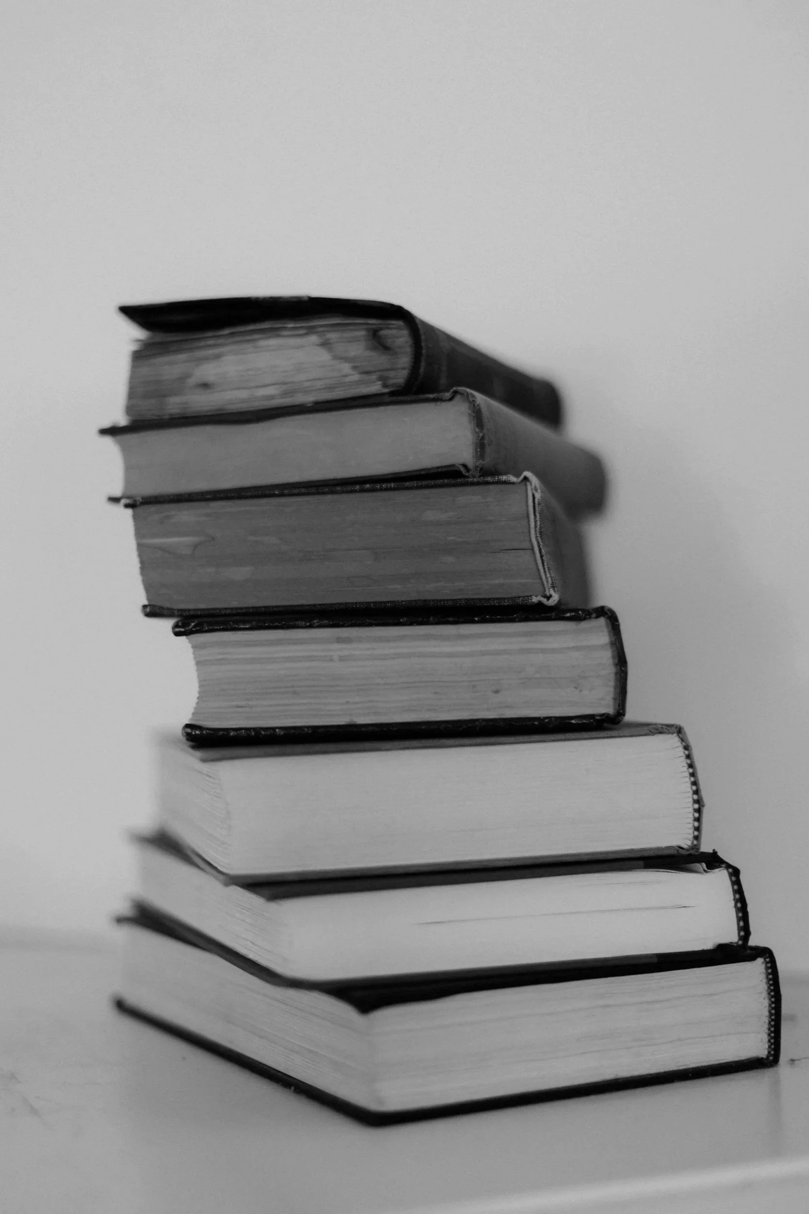 Black and white photo of a neatly stacked pile of books, arranged in a diagonal line on a plain surface.