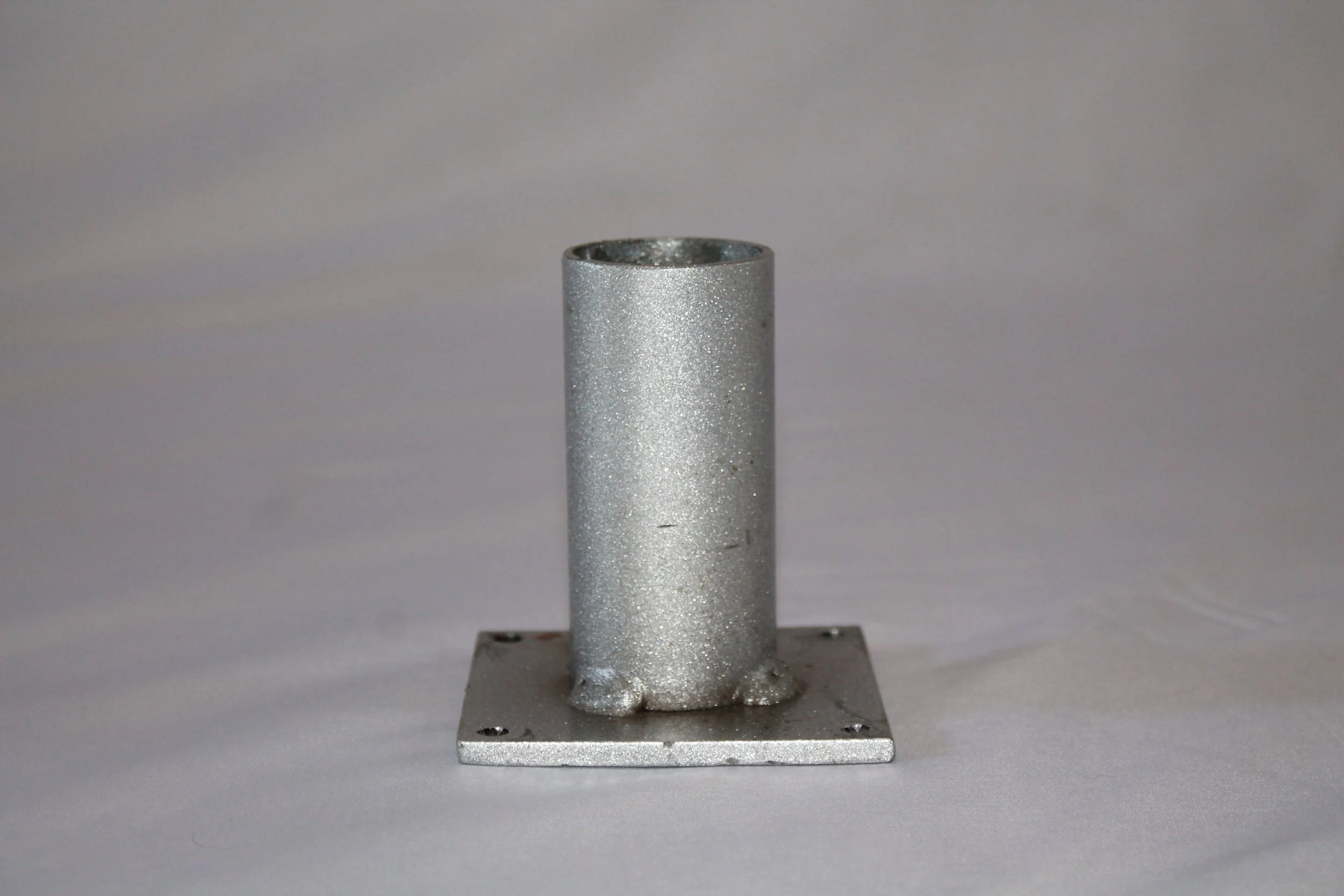 A metallic, cylindrical object mounted on a square metal base with four holes, set against a plain gray background.