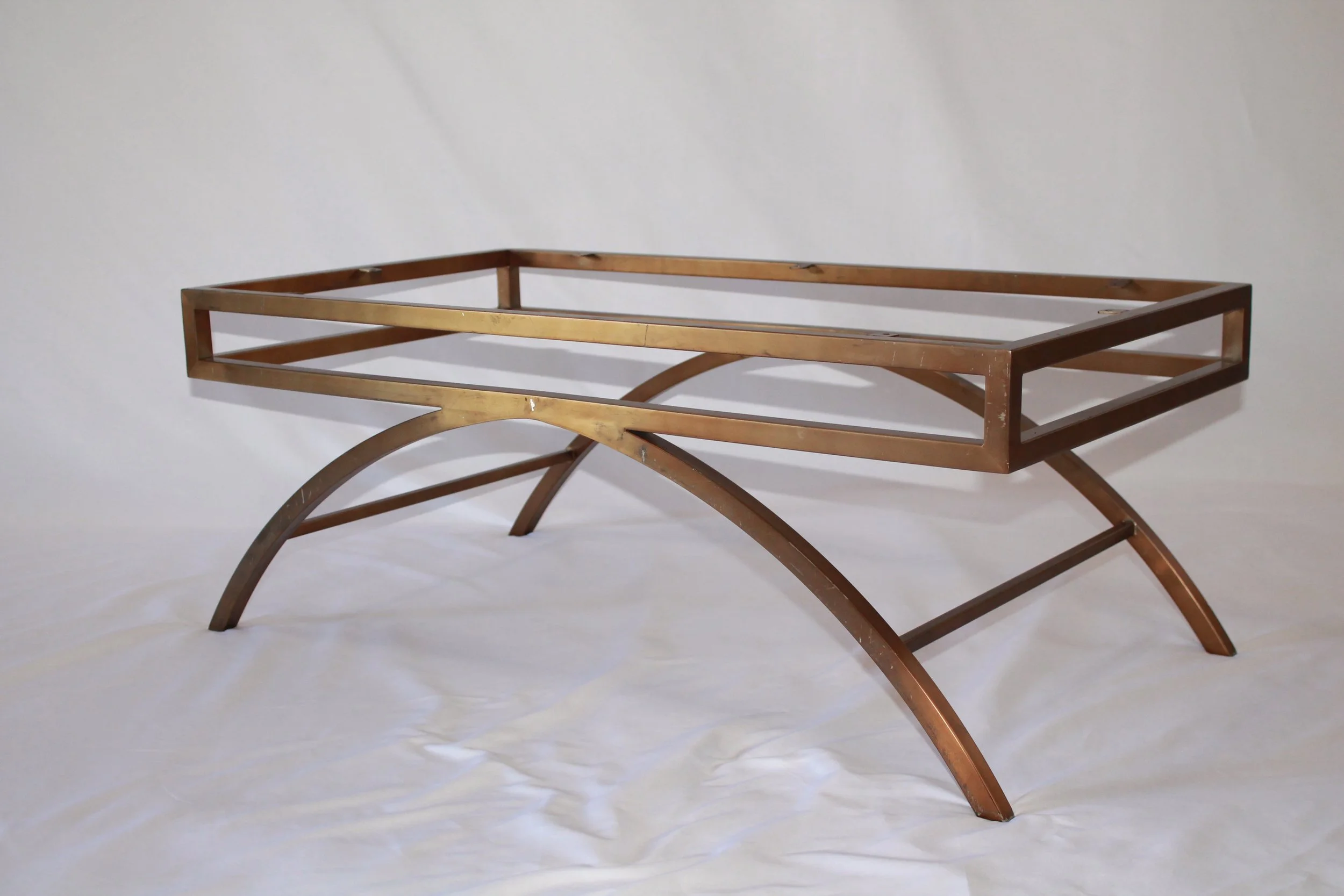 A modern bed frame with a gold-colored metal finish, featuring a rectangular headboard and curved legs, is photographed in a white studio setting.