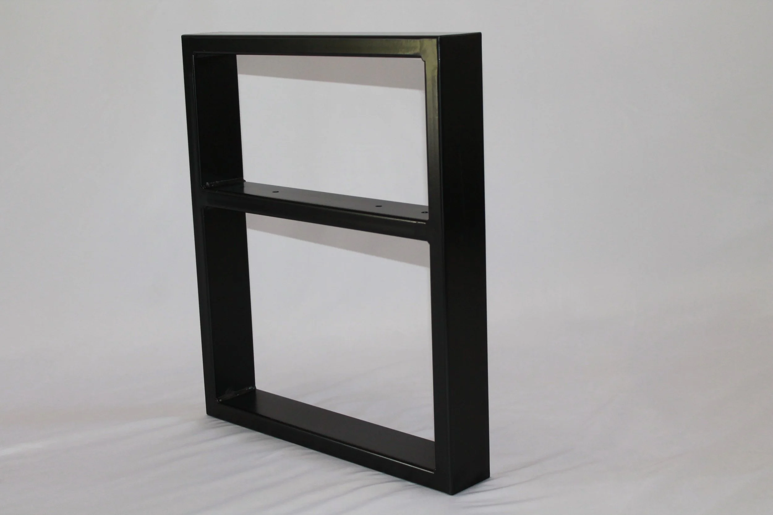 Black rectangular metal frame standing upright on a flat surface, casting a shadow on a plain light-colored background.