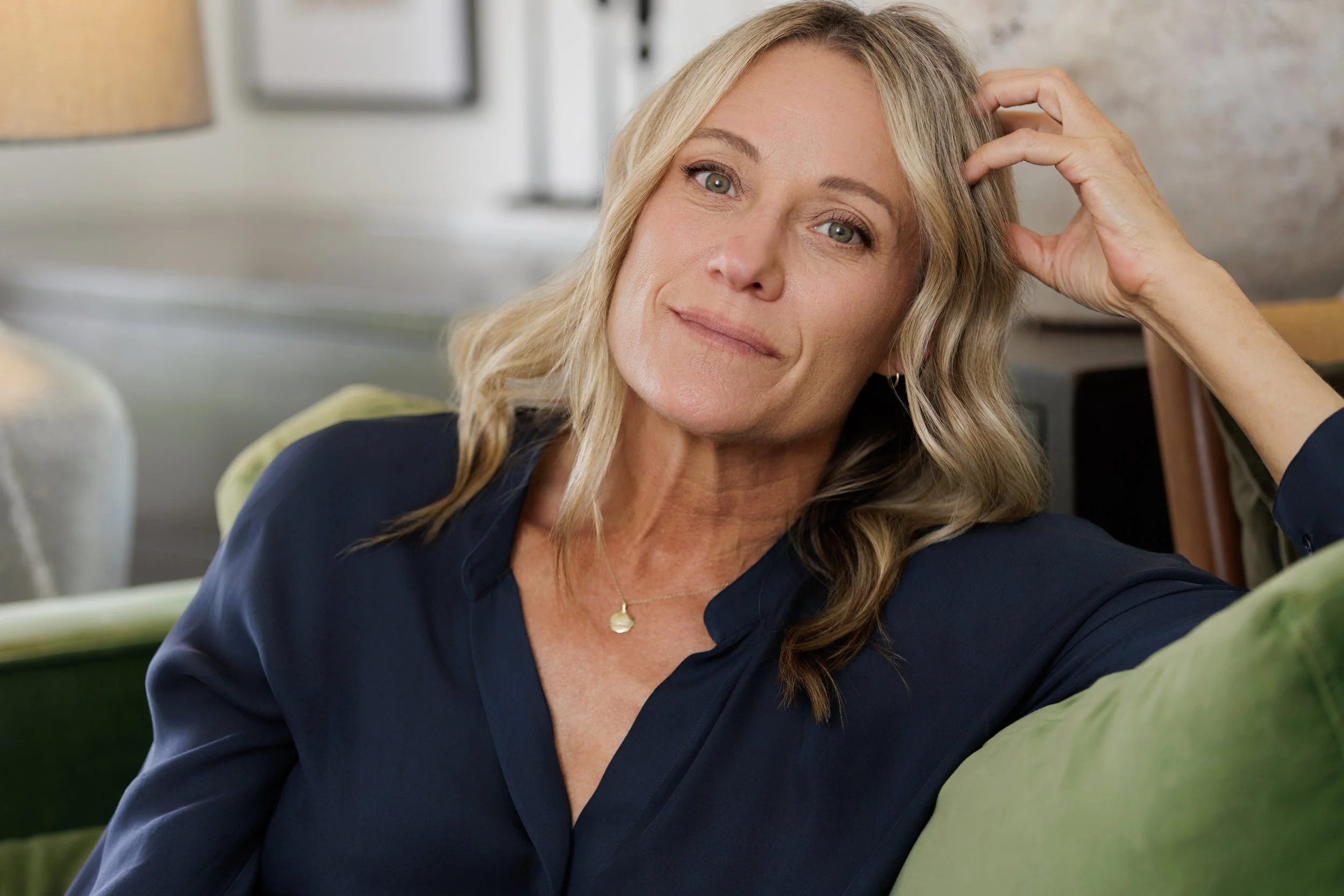A woman with blonde hair and wearing a navy blue top, sitting on a green couch, looking at the camera with her hand touching her hair.