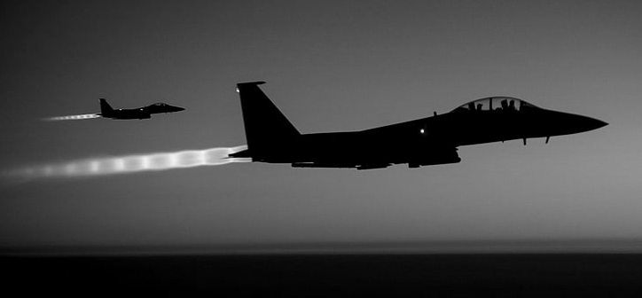 Two jets flying at high speed in formation, with one jet slightly ahead of the other against a dark sky.