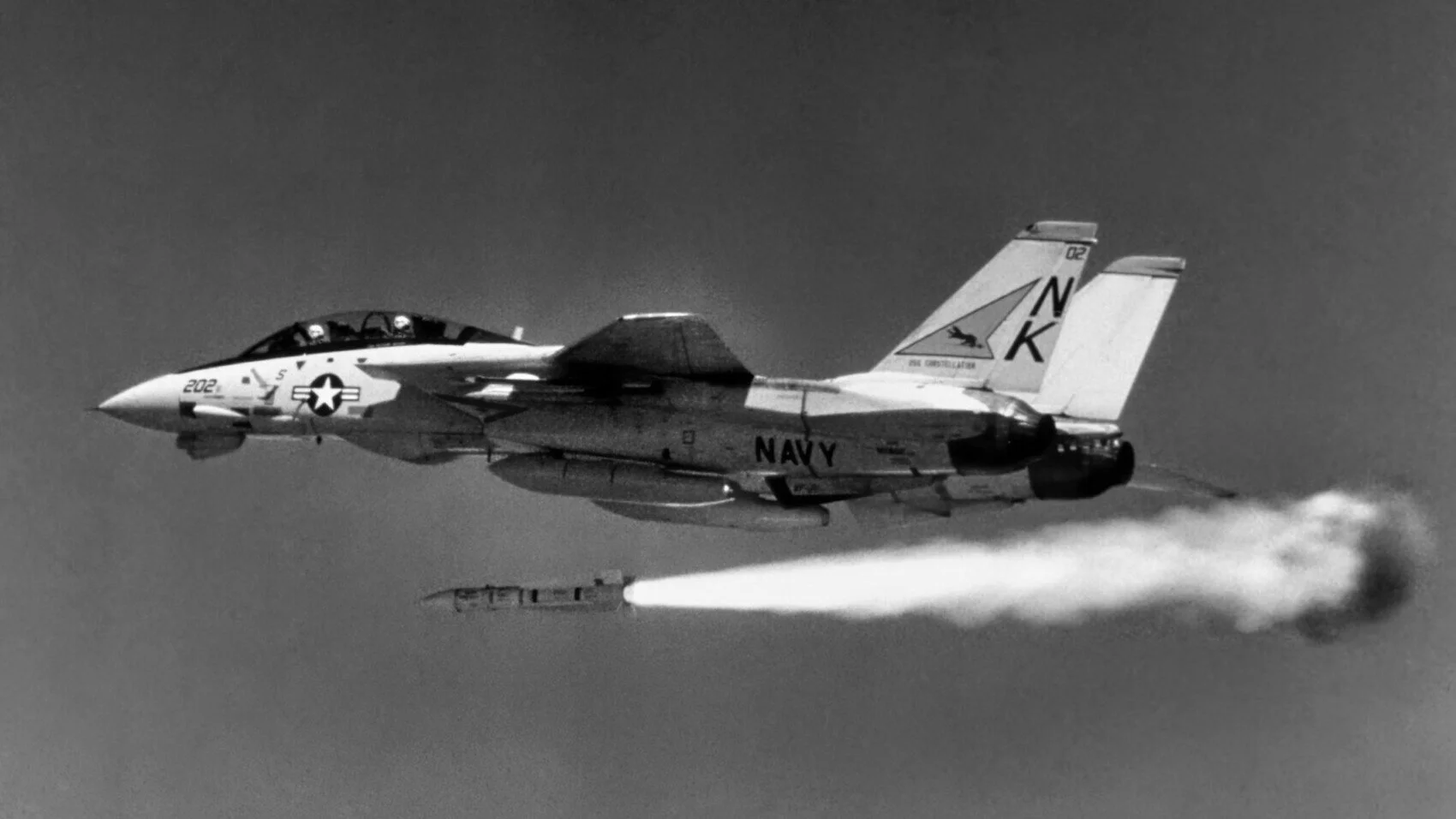 Black and white photo of a US Navy fighter jet flying through the sky, emitting a trail of smoke or vapor.