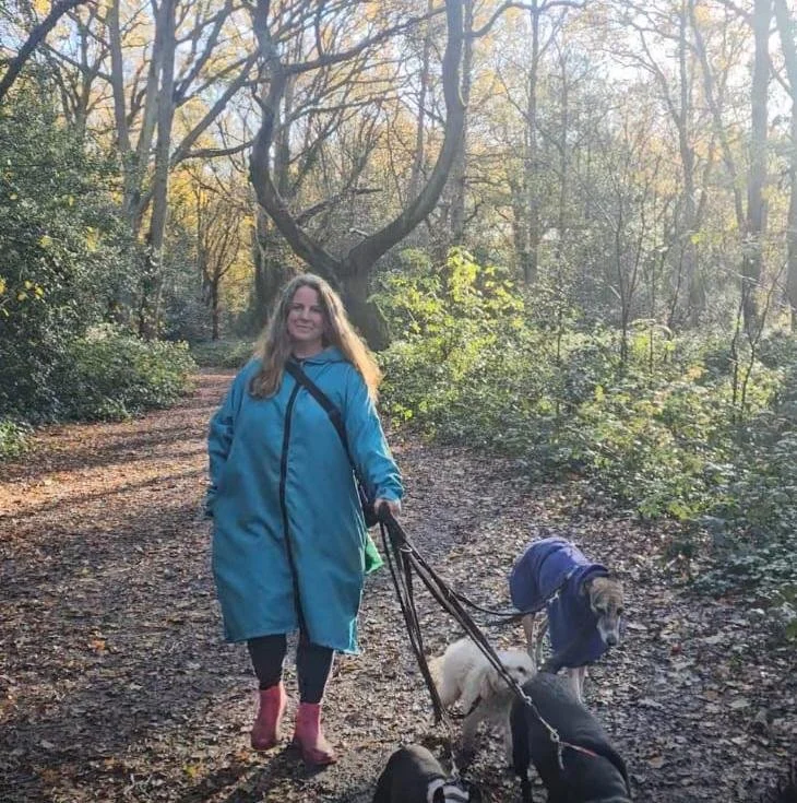 Woman walking in a wooded park with three dogs on a trail during autumn, wearing a blue coat and red boots.