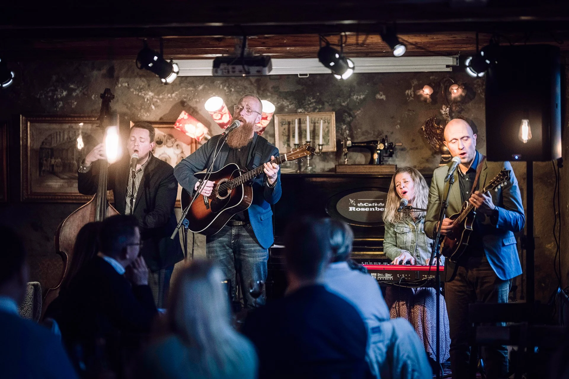 A musical performance at a cozy, dimly lit venue. Four musicians are on stage: a man with glasses and a beard singing and playing an acoustic guitar, a young girl singing into a microphone and playing a keyboard, a man with short dark hair playing an