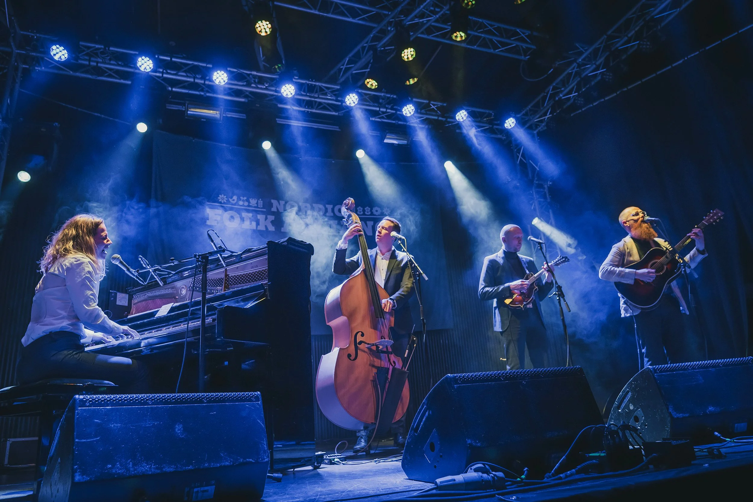 A musical band performing on stage with a pianist, a double bass player, two guitar players, and a vocalist. Stage lighting and a sign with the word 'NORDIC FOLK' are visible in the background.