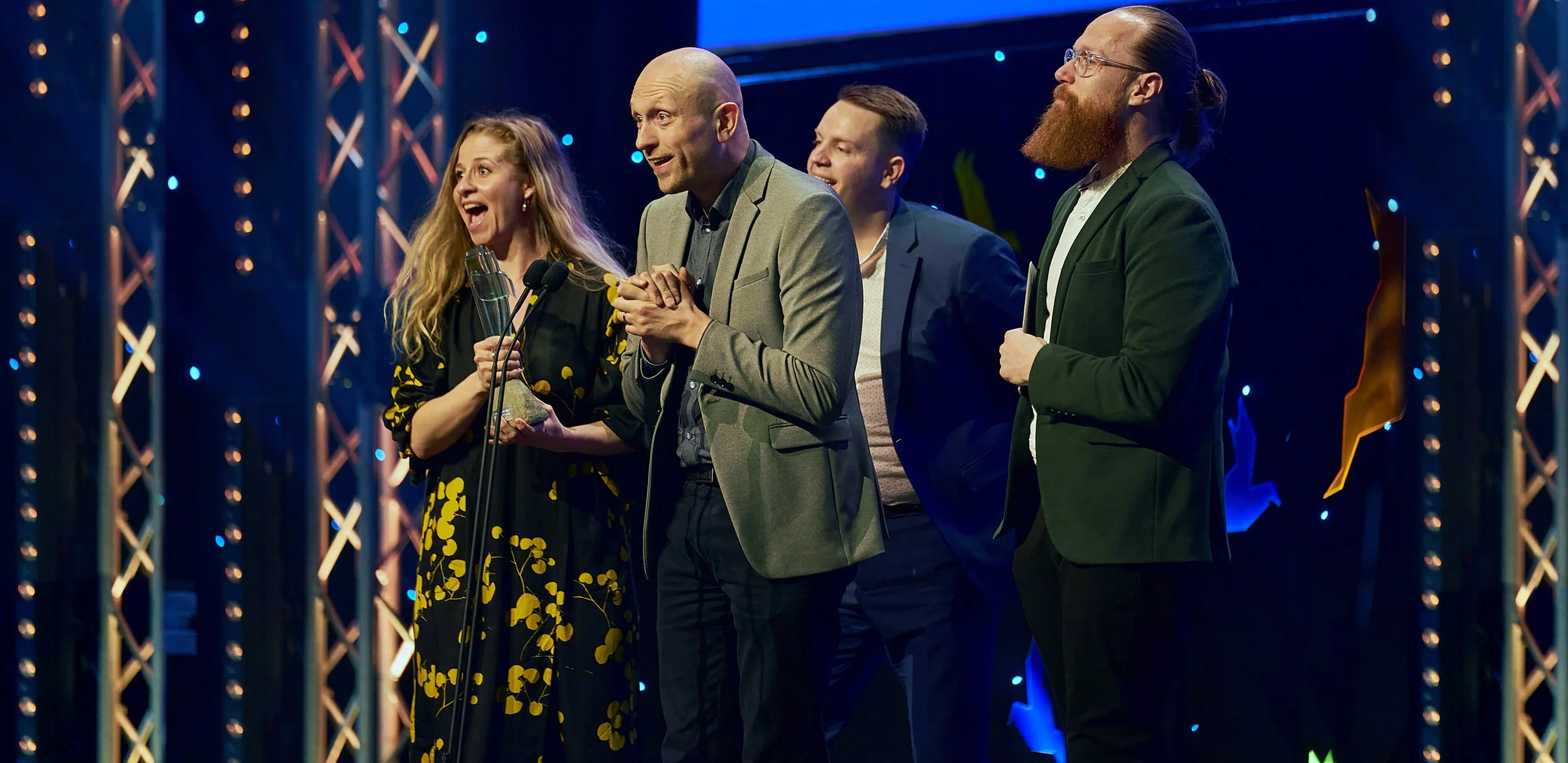 Group of five people on stage, three men and two women, celebrating at an awards ceremony, with one woman holding a trophy, in front of a decorated backdrop with blue lights.