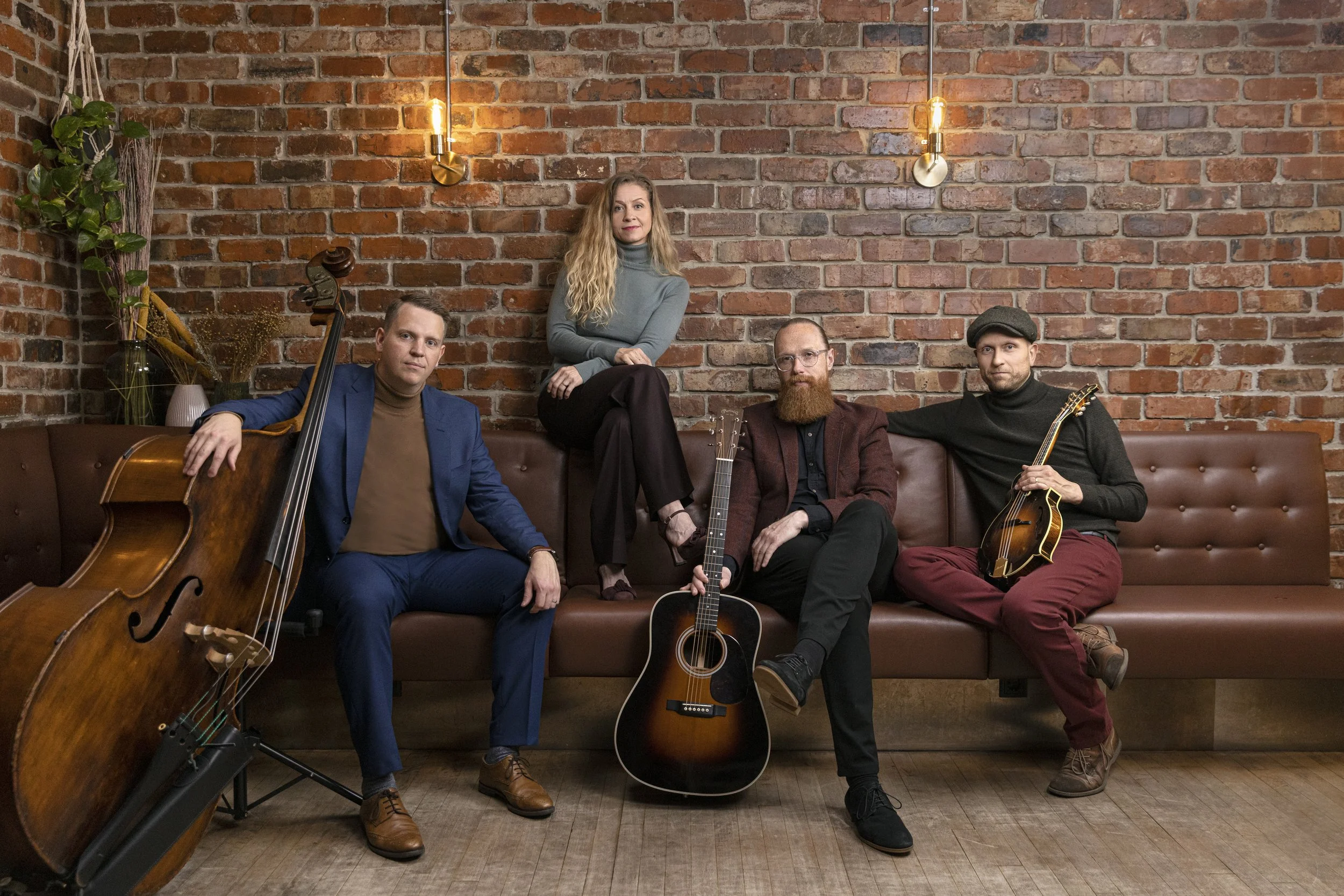 A group of five musicians with guitars and a cello, sitting and standing against a brick wall. The woman in the center is sitting on a ledge, with long blonde hair, wearing a gray turtleneck, and the four men are seated on a brown leather couch, hold