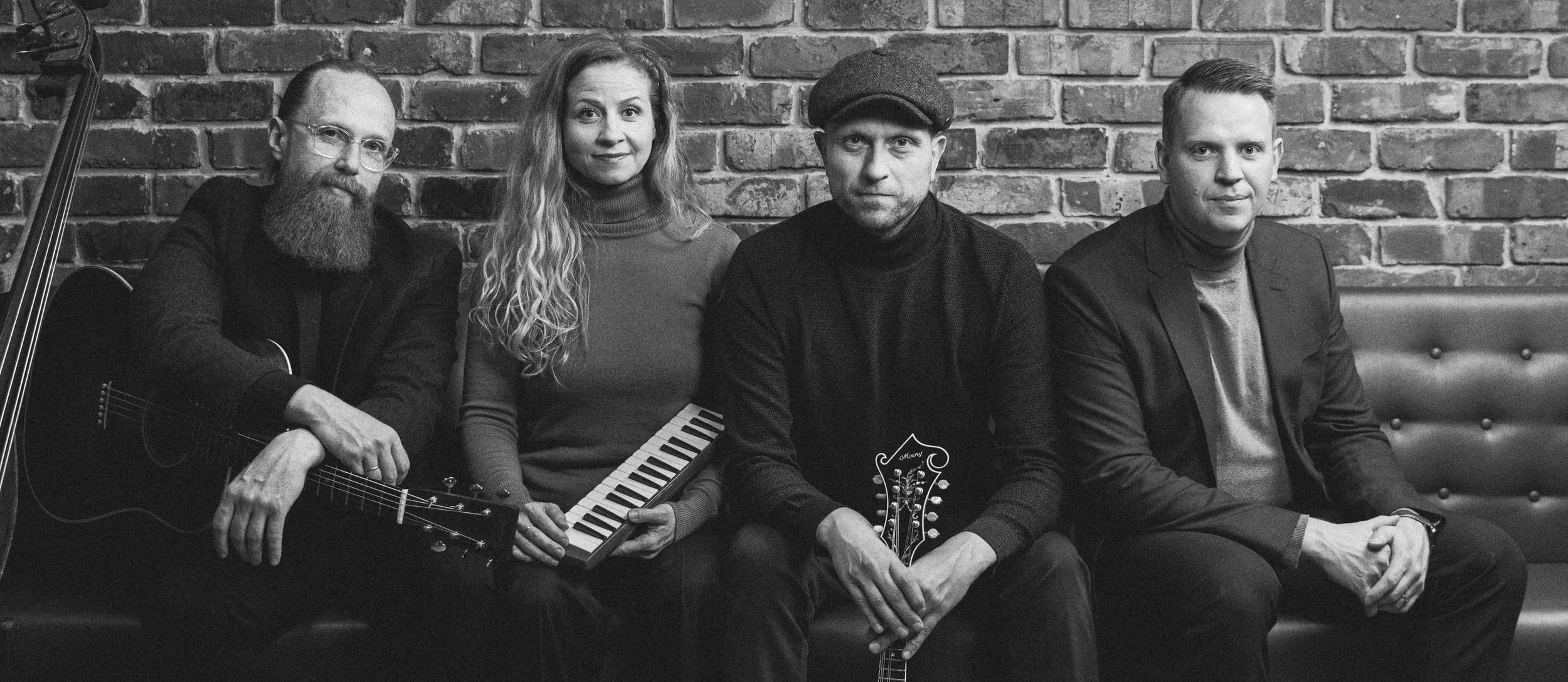 A black and white photo of four musicians sitting on a bench in front of a brick wall. The first man on the left has a beard, glasses, and is holding an acoustic guitar. The woman next to him has long wavy hair and is holding a small keyboard. The third person, a man wearing a flat cap, is holding a ukulele. The fourth person, a man in a blazer, has short hair and is sitting with his hands clasped.