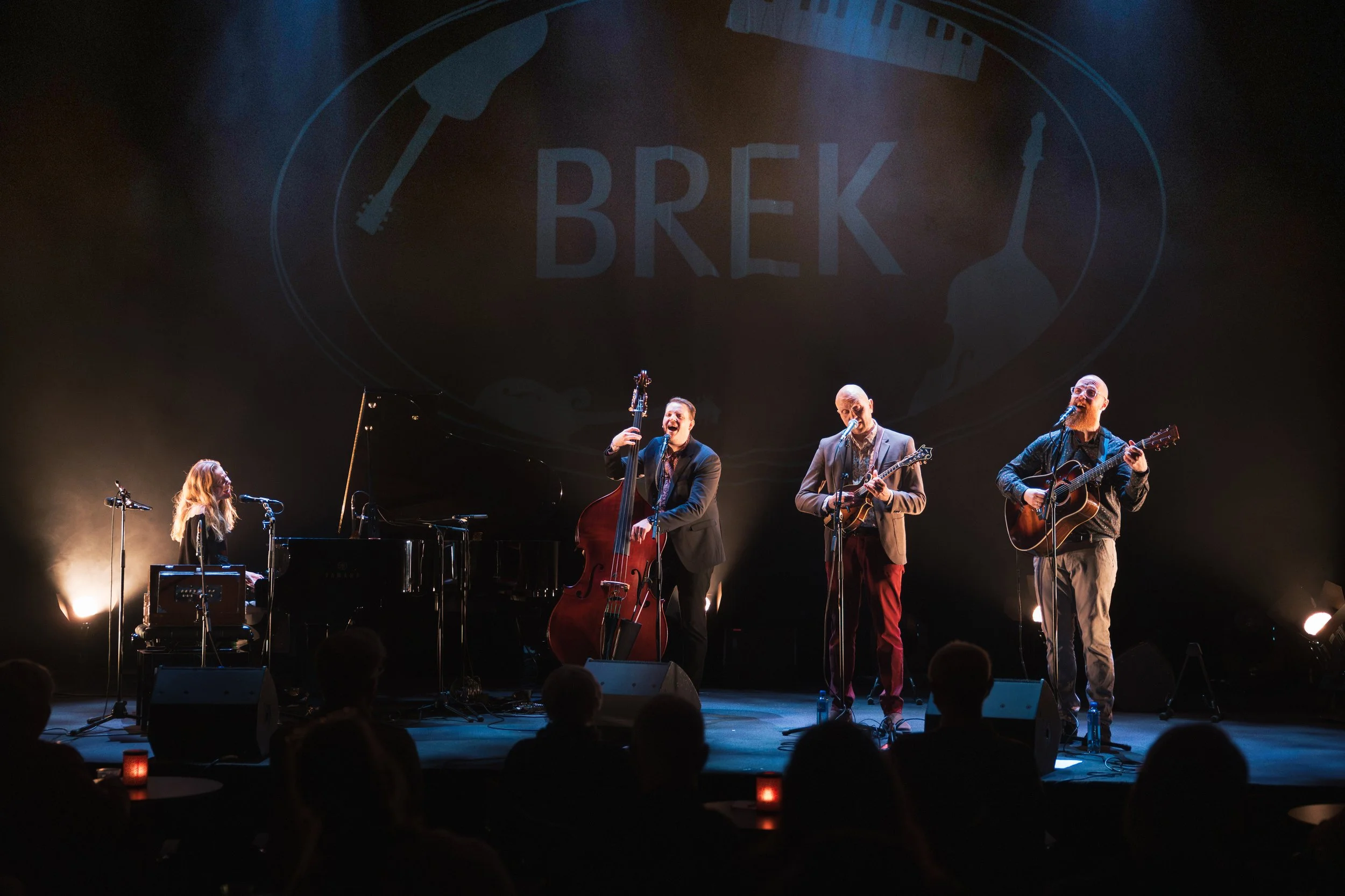A musical performance on stage with five musicians, including a pianist and four guitar players, under a large screen displaying the blue logo 'BREK' with musical instruments around it.