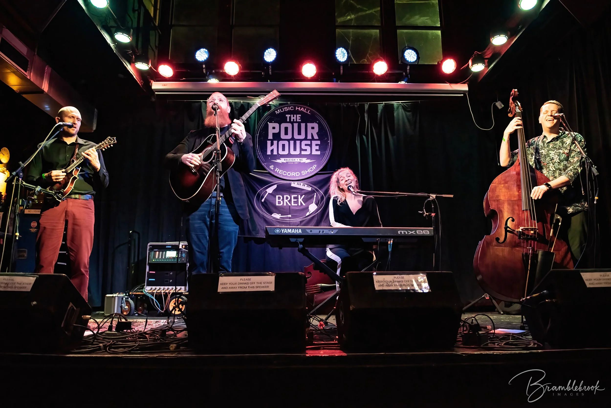 Four musicians performing on stage at The Pour House music hall with a sign behind them, two playing guitars, one singing, and one on keyboard.