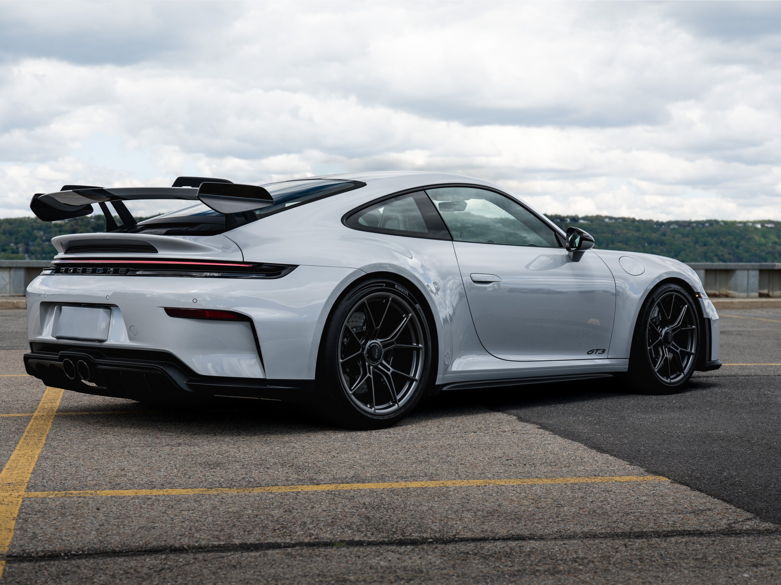 Ceramic gray Porsche 911 GT3 parked in an outdoor lot under cloudy skies with a scenic distant landscape.