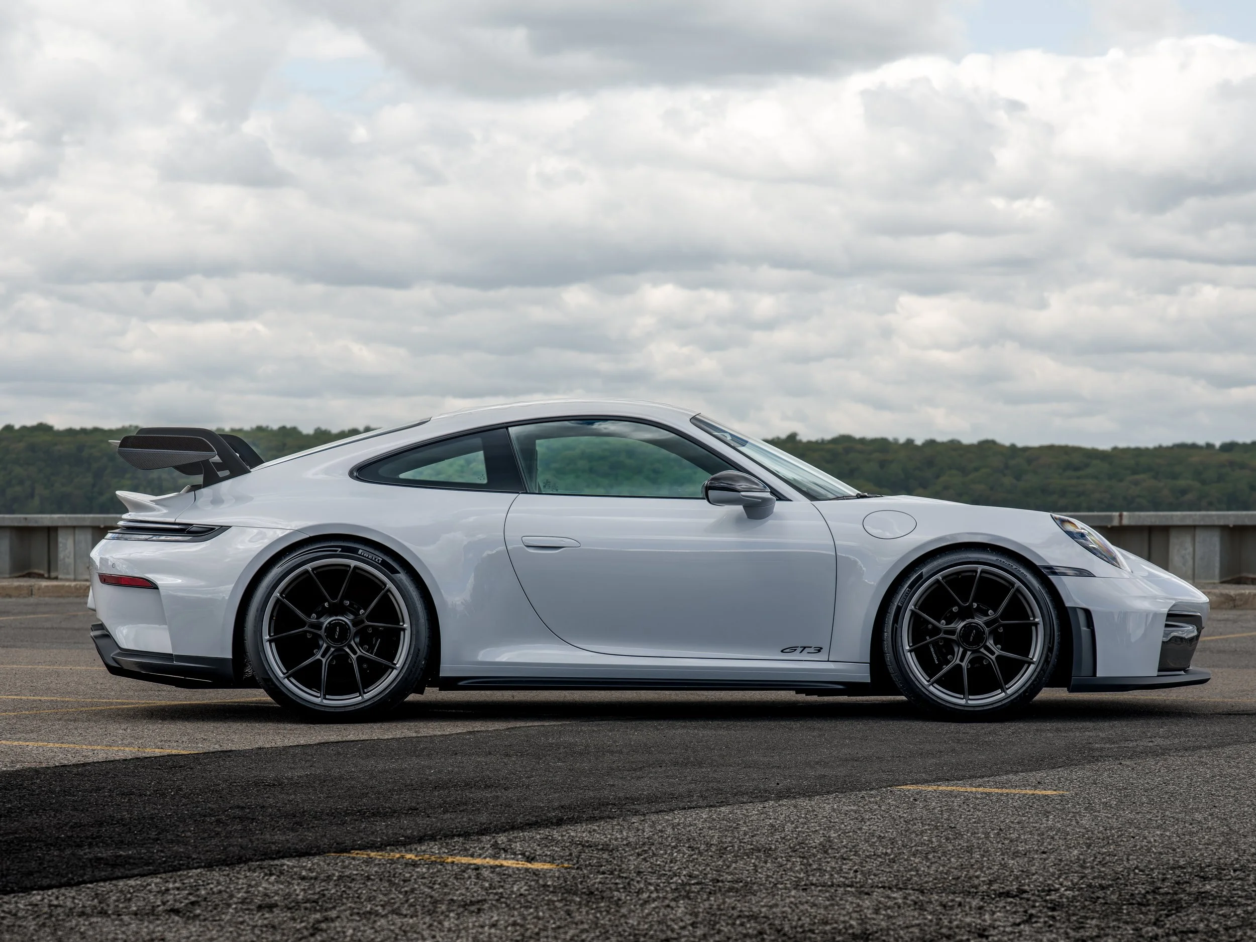 A ceramic gray Porsche 911 GT3 with a rear spoiler and black wheels parked on a paved lot under a cloudy sky.