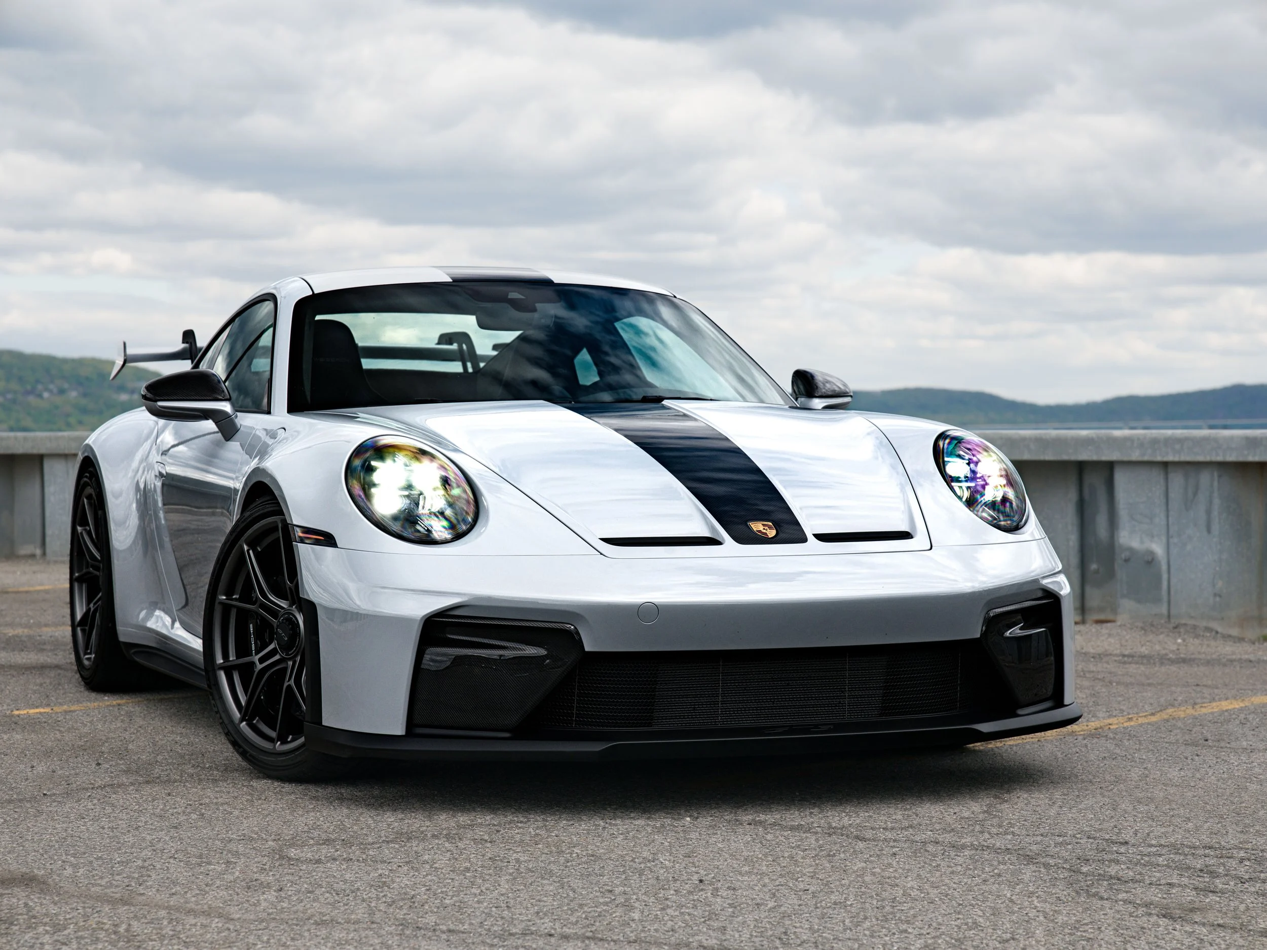 A ceramic gray Porsche 911 GT3 with carbon fiber racing stripe on the hood, parked outdoors on a cloudy day.