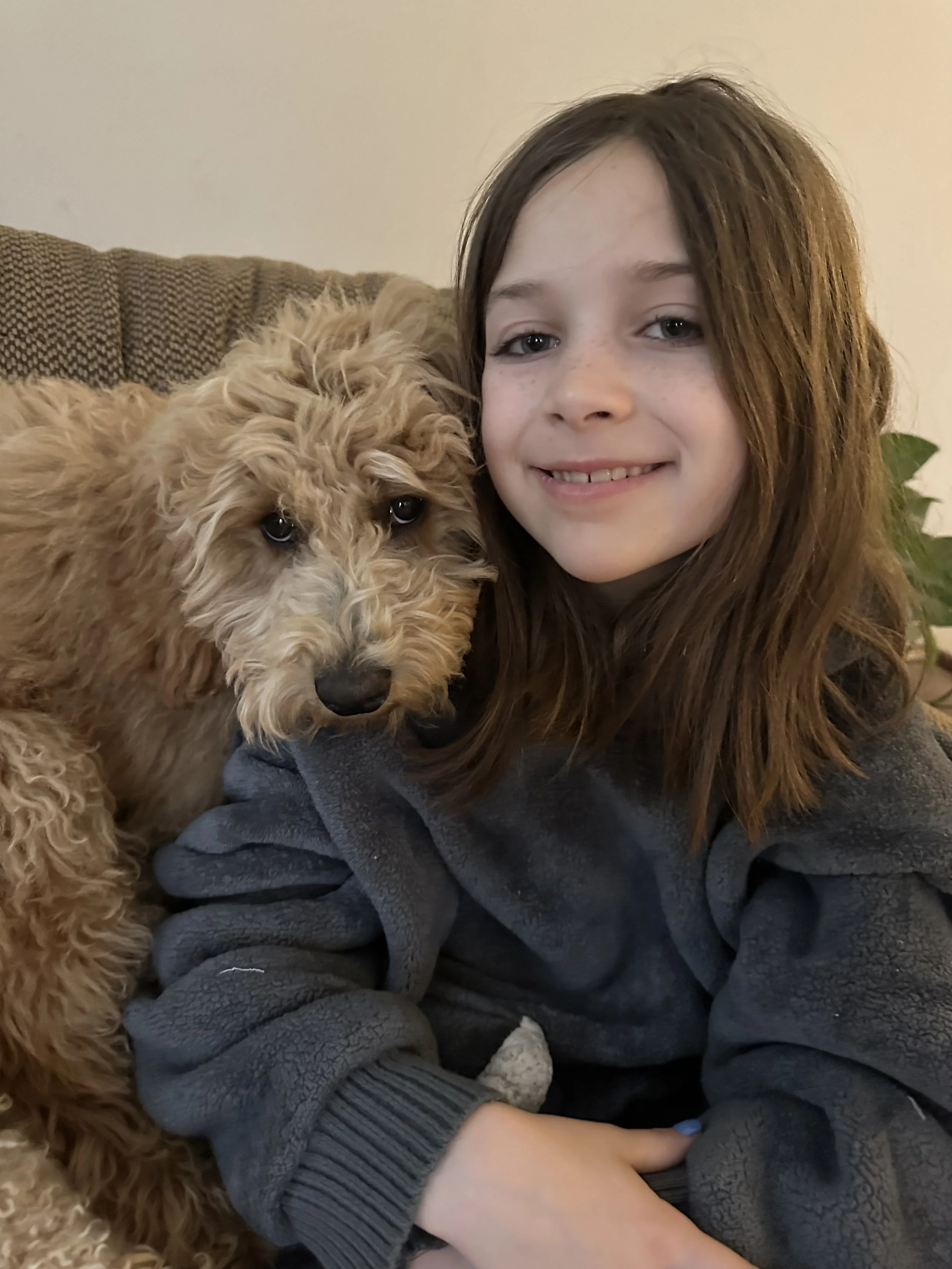 A girl with long reddish-brown hair smiling and sitting on a couch with a curly-haired dog, possibly a Goldendoodle, resting against her.
