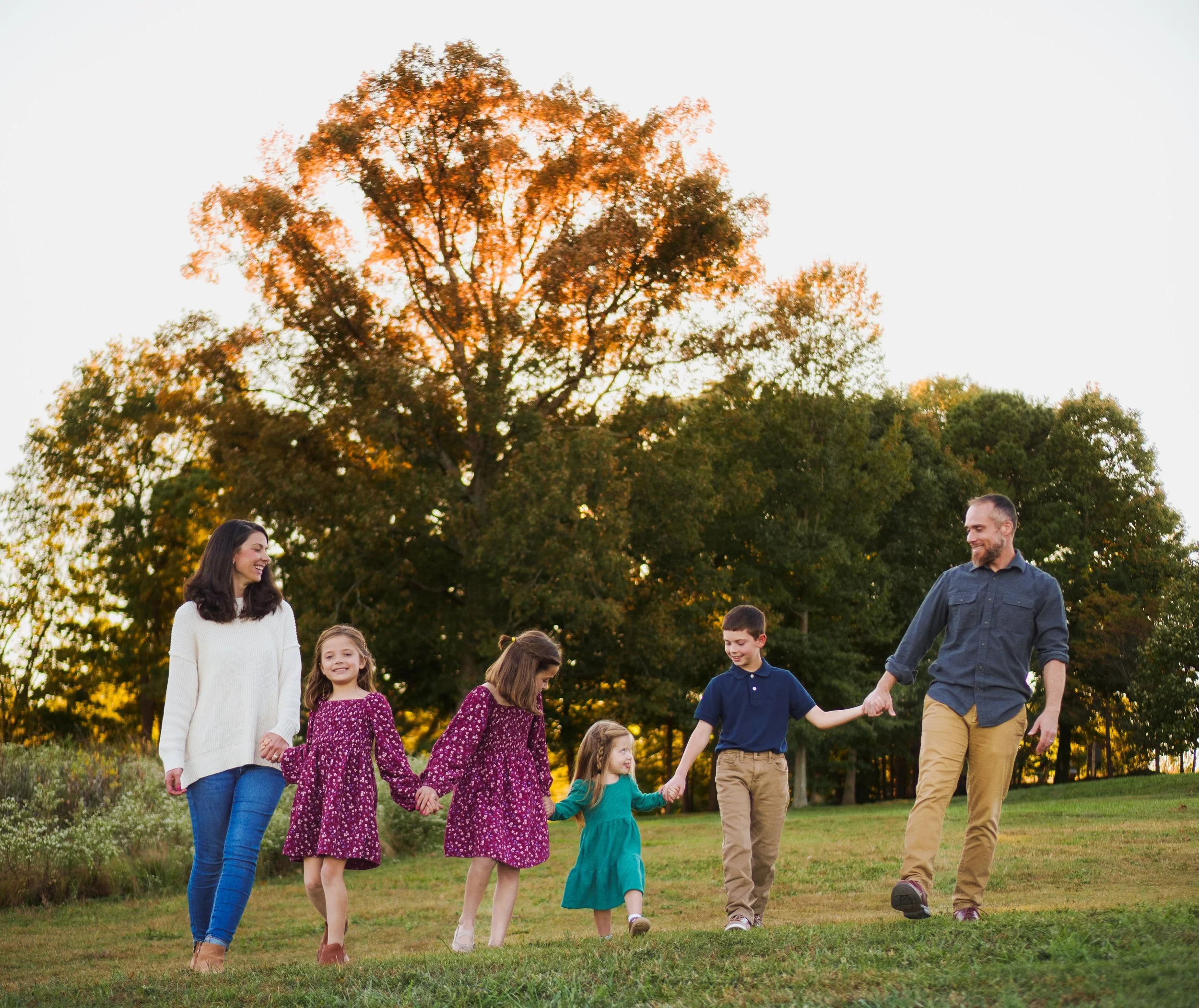 A family walking hand in hand outdoors in a park during autumn, with a large tree with orange leaves in the background.