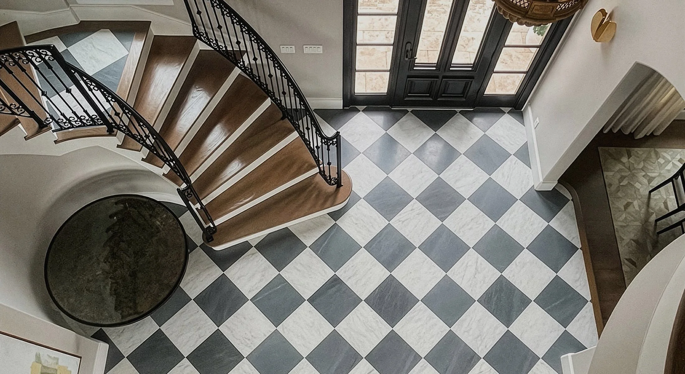 A house entryway with a black front door and glass panels, a checkered black and white tile floor, a wooden staircase with a decorative black metal railing, and a large round black table.