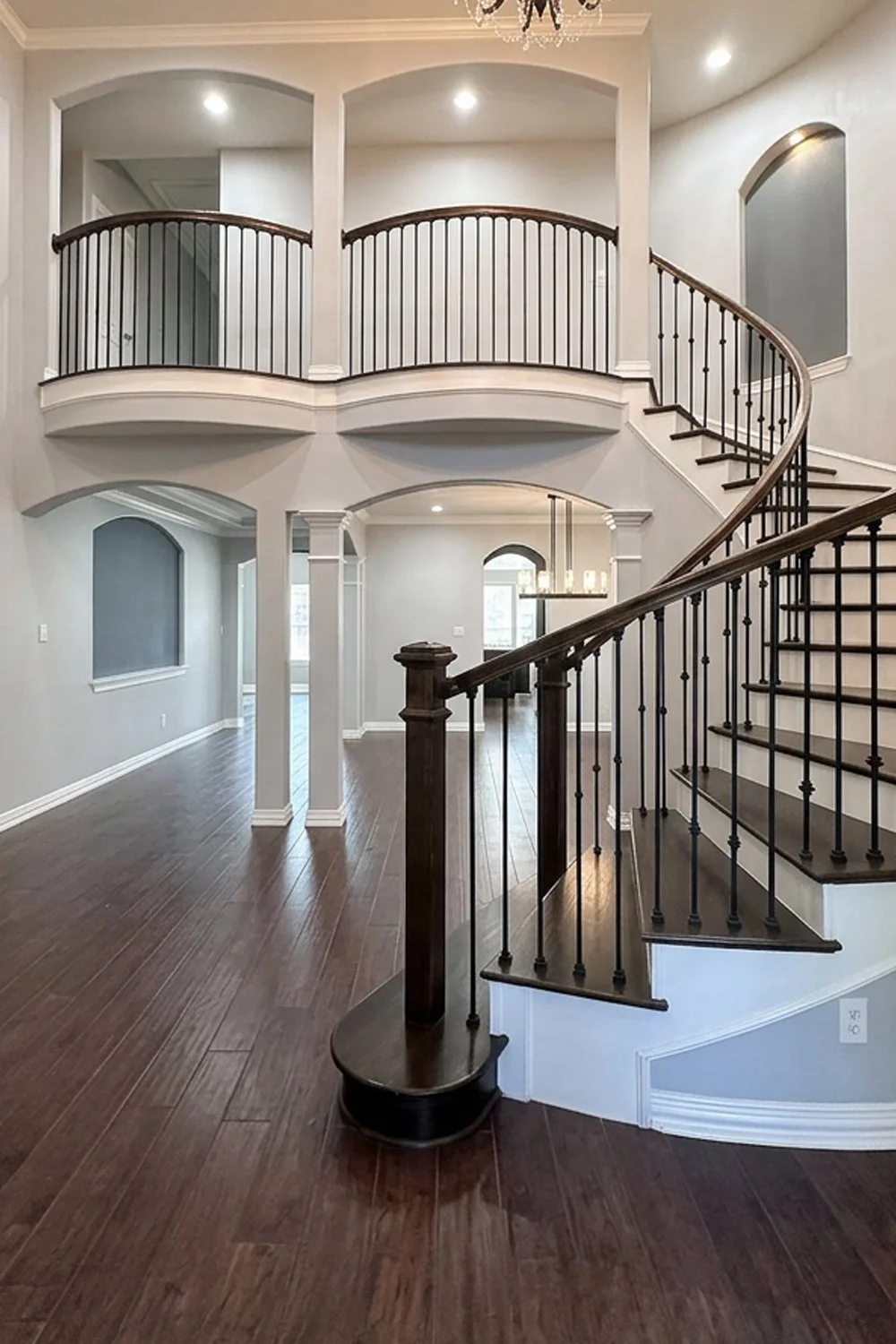 Interior view of a two-story house with dark wood flooring, a curved staircase with wooden steps and dated railings, and a second-floor balcony overlooking the entryway.