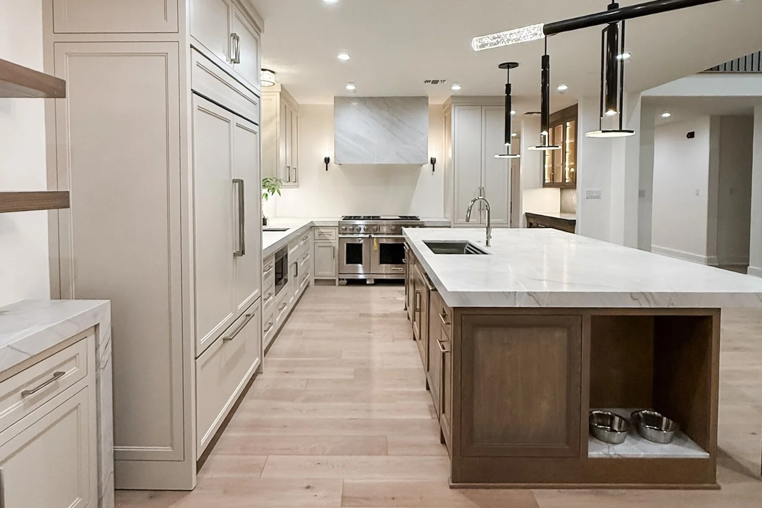 A light, modern kitchen with white cabinets, a large island with a marble countertop, professional appliances, and light oak wood flooring.